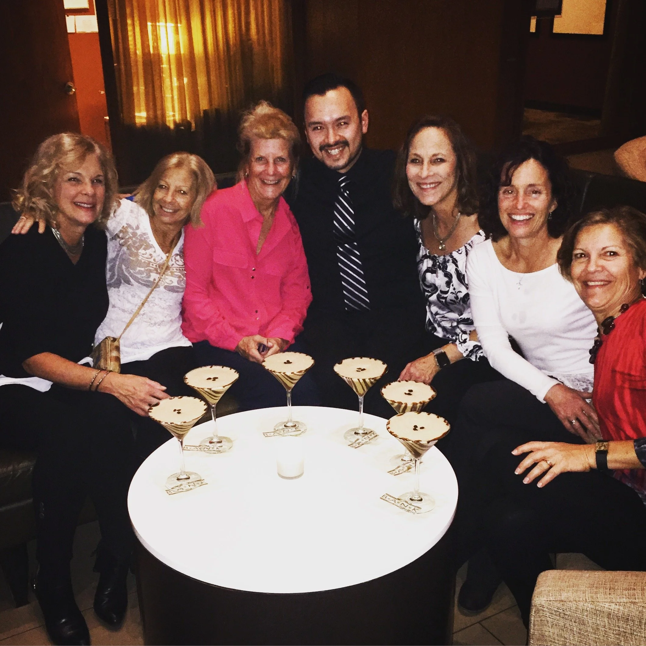 Group of seven people, six women and one man, sitting together at a round table with eight cocktails with martini glasses, in a dimly lit room, smiling at the camera.