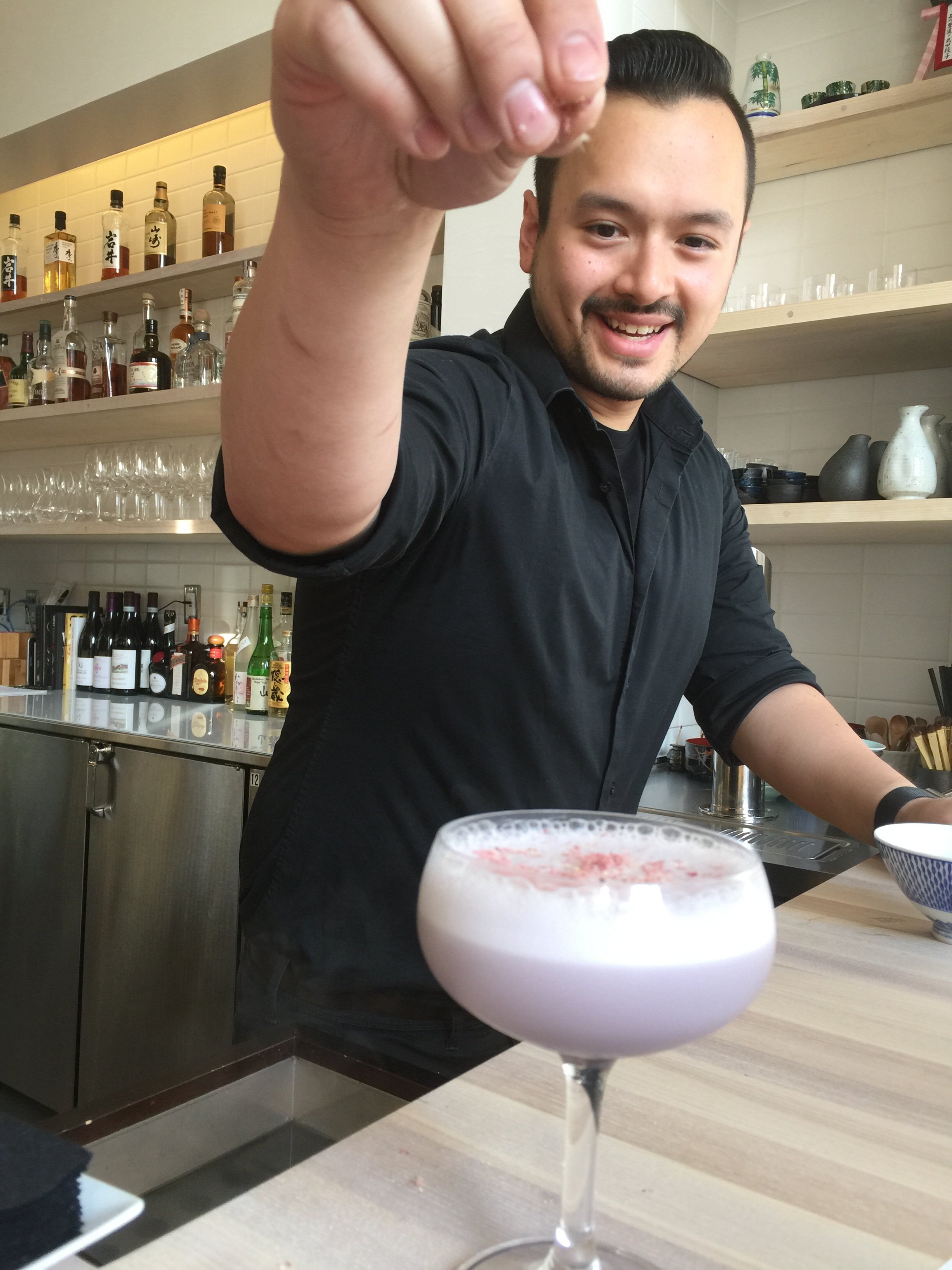 A smiling bartender in black shirt reaching towards the camera with a cocktail in a coupe glass in the foreground. The bar has bottles of liquor and glassware on shelves behind him.
