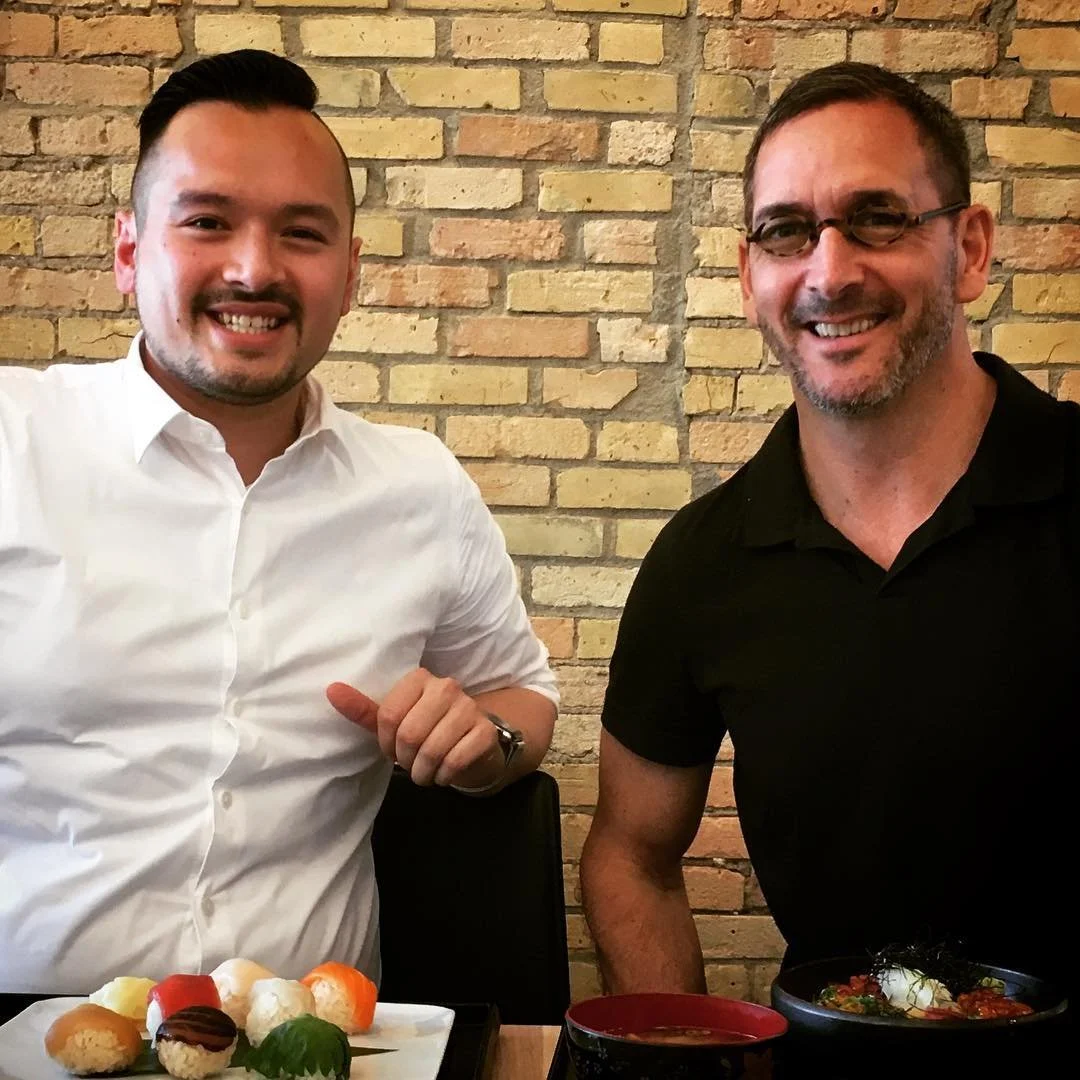 Two men sitting at a table in a restaurant, smiling, with sushi and dishes in front of them, against a brick wall background.