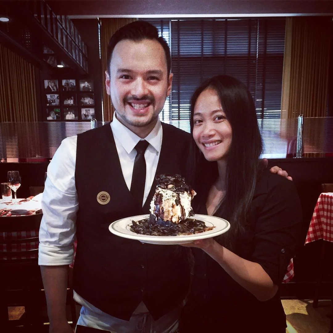 A man and a woman in a restaurant holding a plate with a large dessert topped with ice cream, chocolate syrup, and cookies.