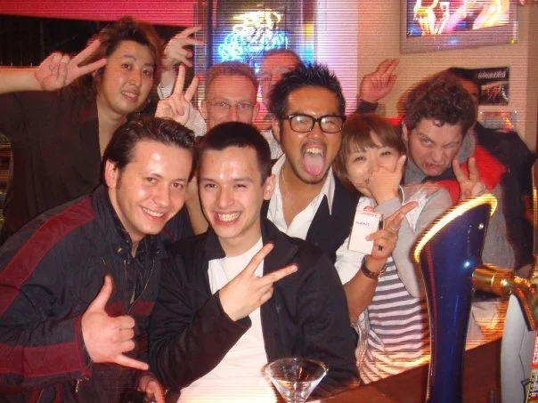 Group of diverse friends at a bar or restaurant, smiling and making peace signs and gestures, with neon lights in the background.