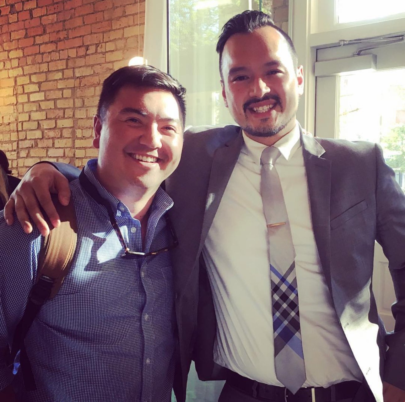 Two men smiling, one wearing a backpack, and the other in a suit with a plaid tie, standing inside near a large window with sunlight coming through.