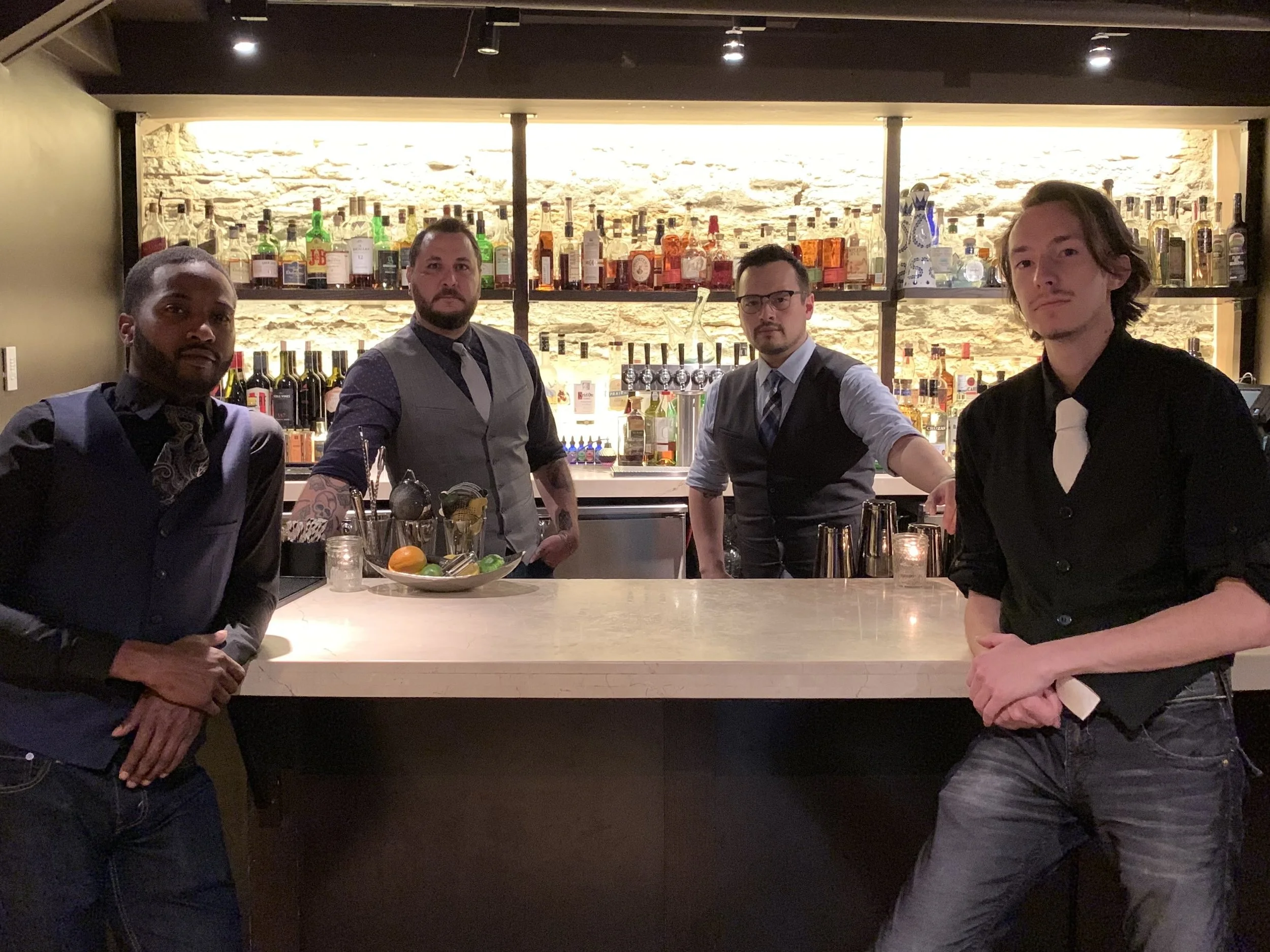 Four bartenders in formal attire behind a bar with bottles of alcohol on shelves in a dimly lit bar setting.