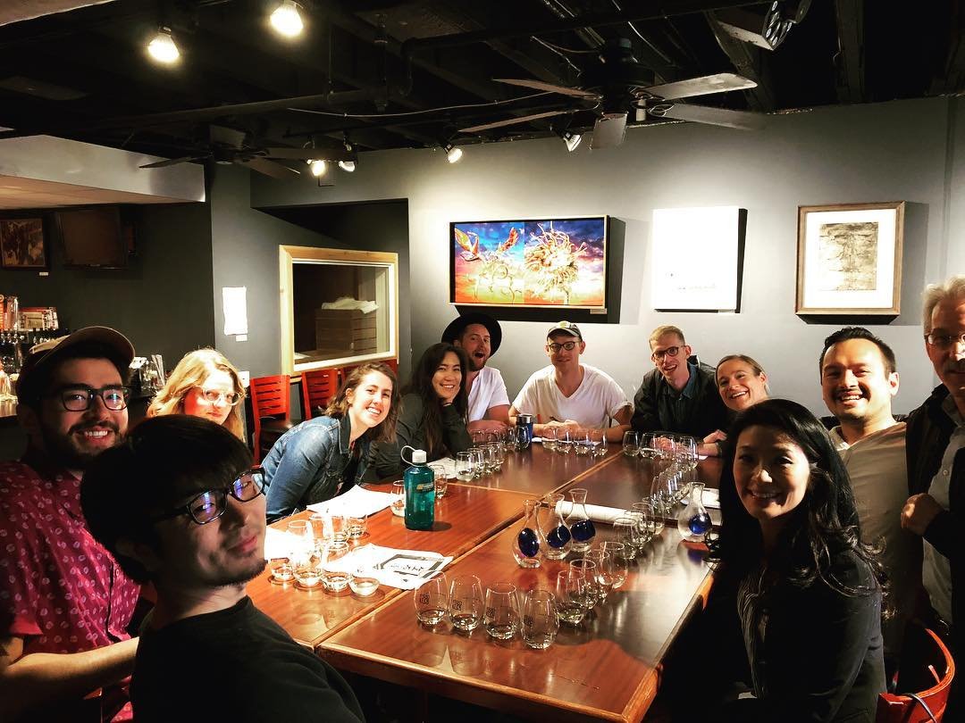 A group of twelve people sitting around a banquet table in a dimly lit restaurant or bar, smiling at the camera, with glasses and water bottles on the table, and artwork on the wall behind them.