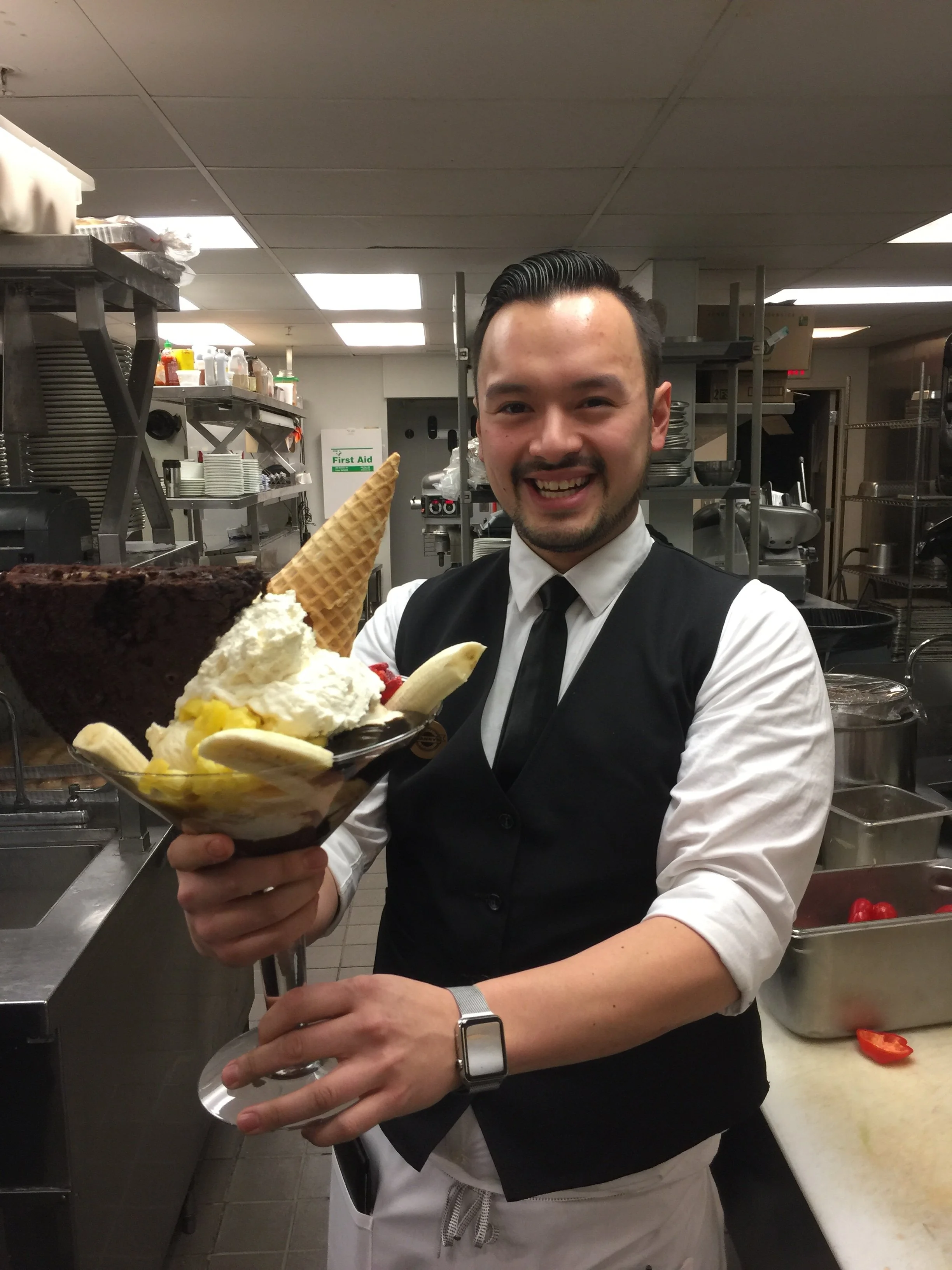 A smiling man dressed in a black vest, white shirt, and black tie holding a large bowl of ice cream with various toppings and garnishes in a commercial kitchen.