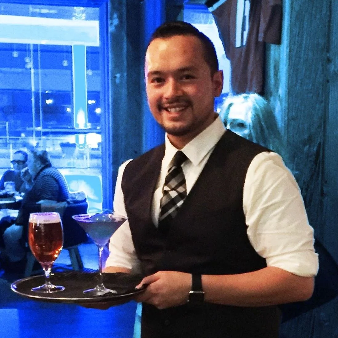 A smiling man in a white shirt, black vest, and plaid tie holding a tray with a glass of red wine, a cocktail, and a martini in a dimly lit restaurant or bar with blue-tinted lighting.