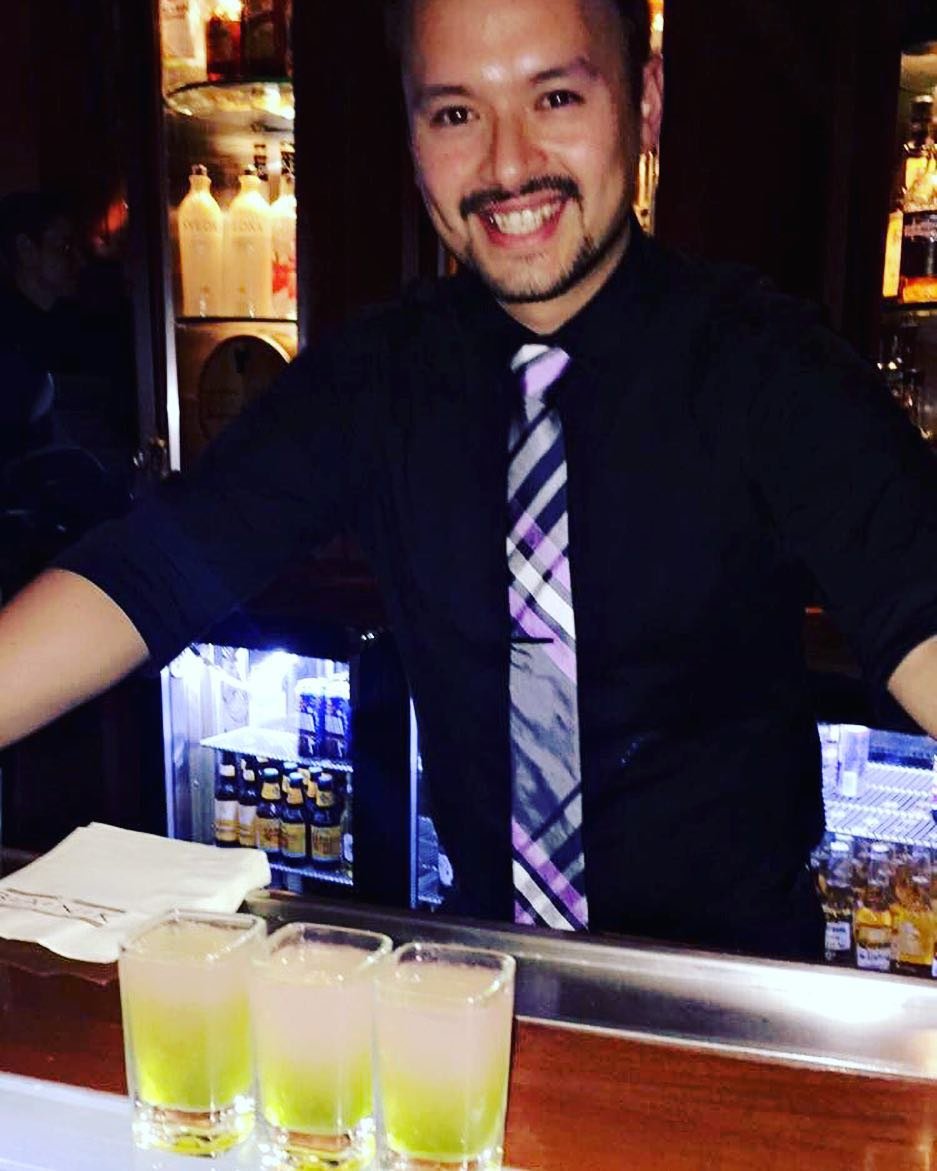A bartender with a beard smiling behind the bar, with three glasses containing a yellow-green drink in front of him, in a dimly lit bar or pub.