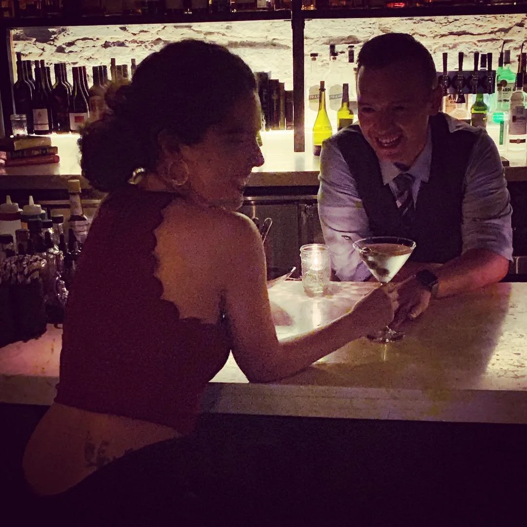 A woman and a bartender sharing a laugh at a bar, with the woman holding a cocktail.