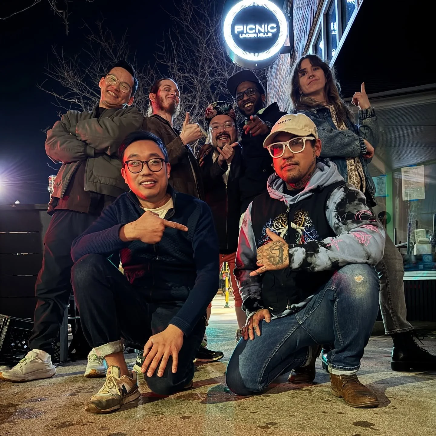 Group of nine diverse friends posing outside at night in front of a lit sign that reads 'Picnic Union Hall.' They are smiling, making gestures, and dressed casually.