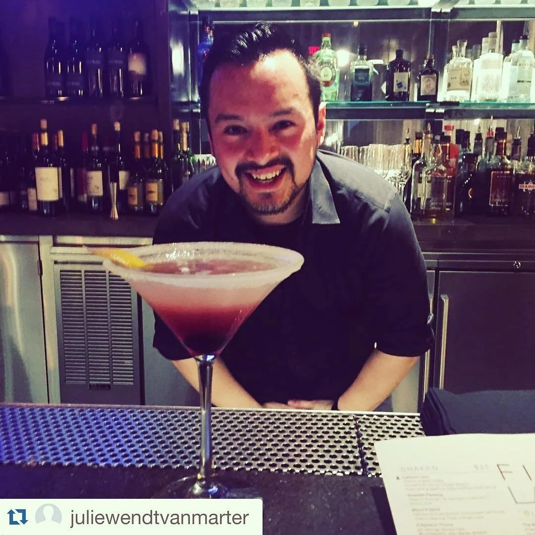 A bartender smiling behind a bar counter with a pink cocktail garnished with a lemon slice in the foreground.