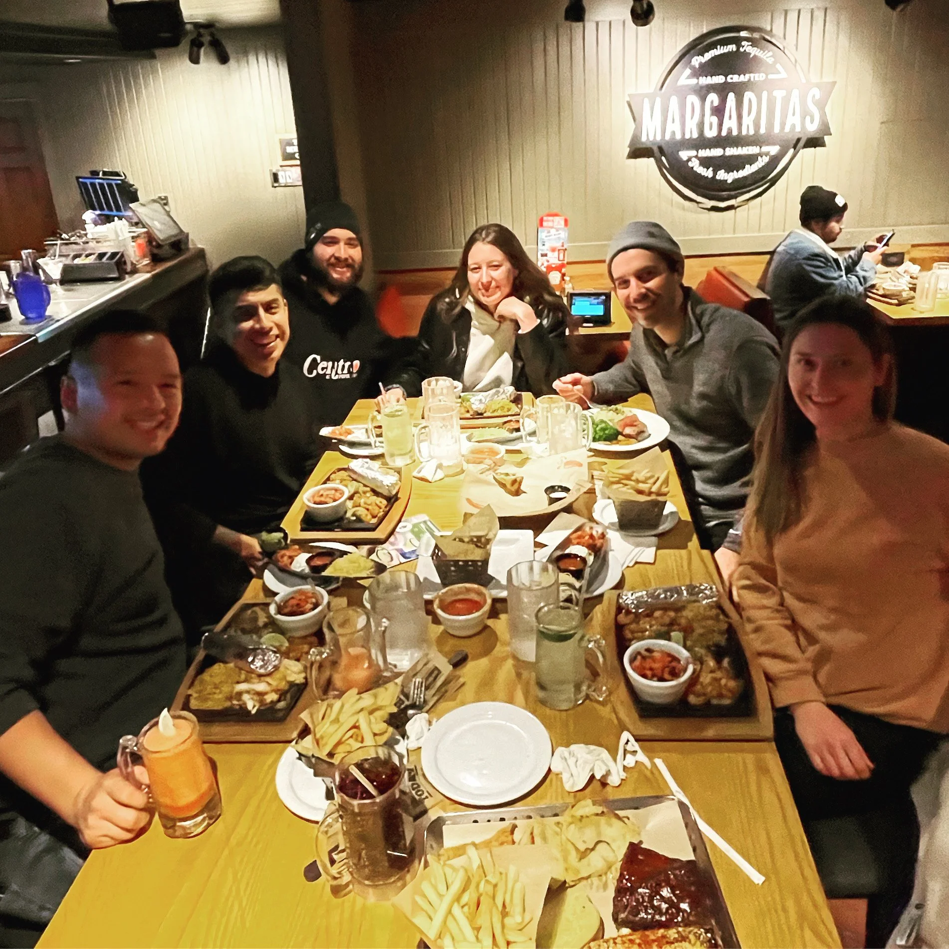 Group of six friends sitting at a restaurant table enjoying Mexican food, with drinks and plates of food including tacos, nachos, and bowls of salsa and beans.
