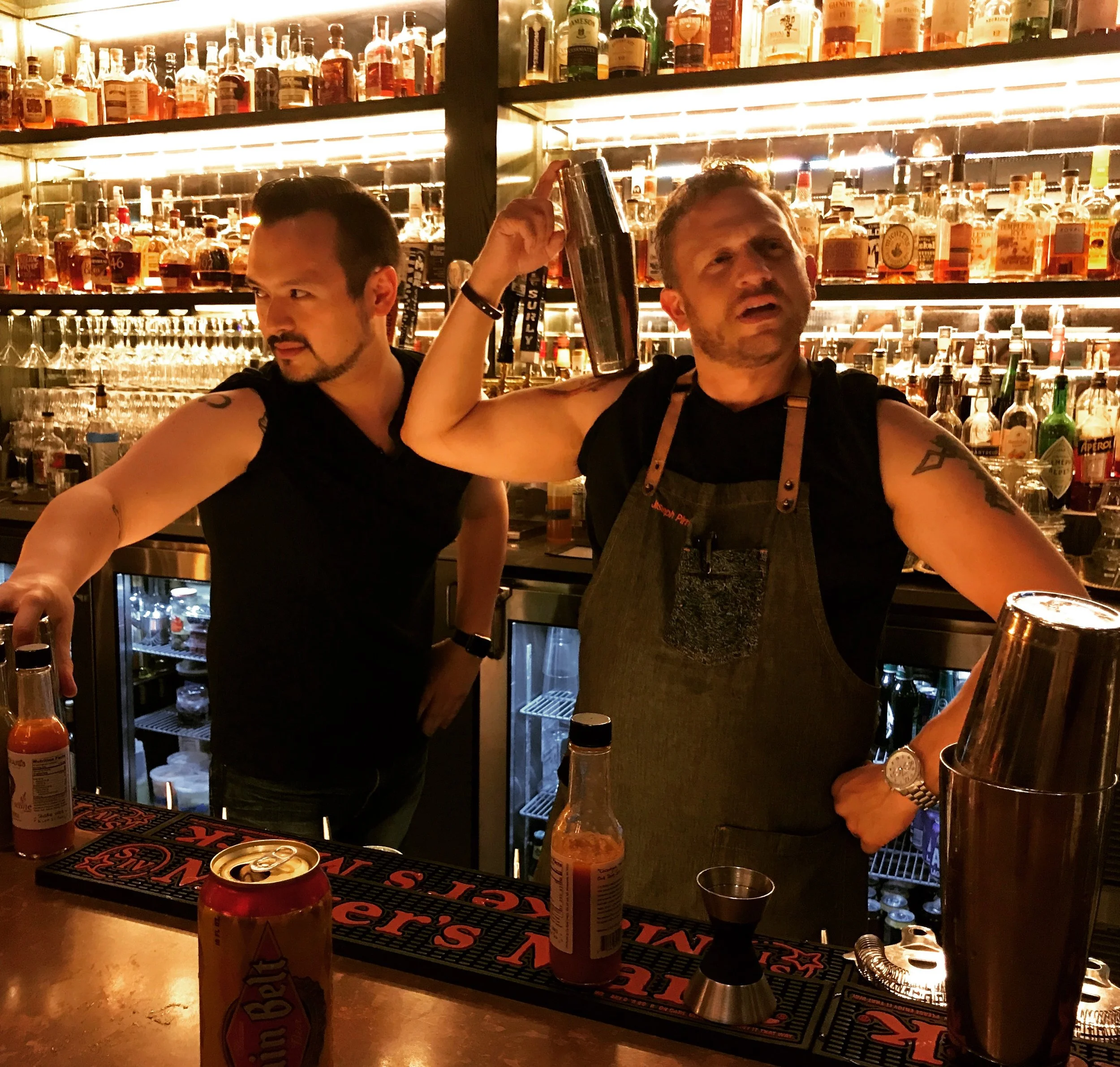 Two bartenders behind a bar with a collection of liquor bottles on shelves in the background. One bartender is a man with tattoos, wearing a dark sleeveless shirt and an apron, holding a cocktail shaker on his shoulder. The other bartender is a man w