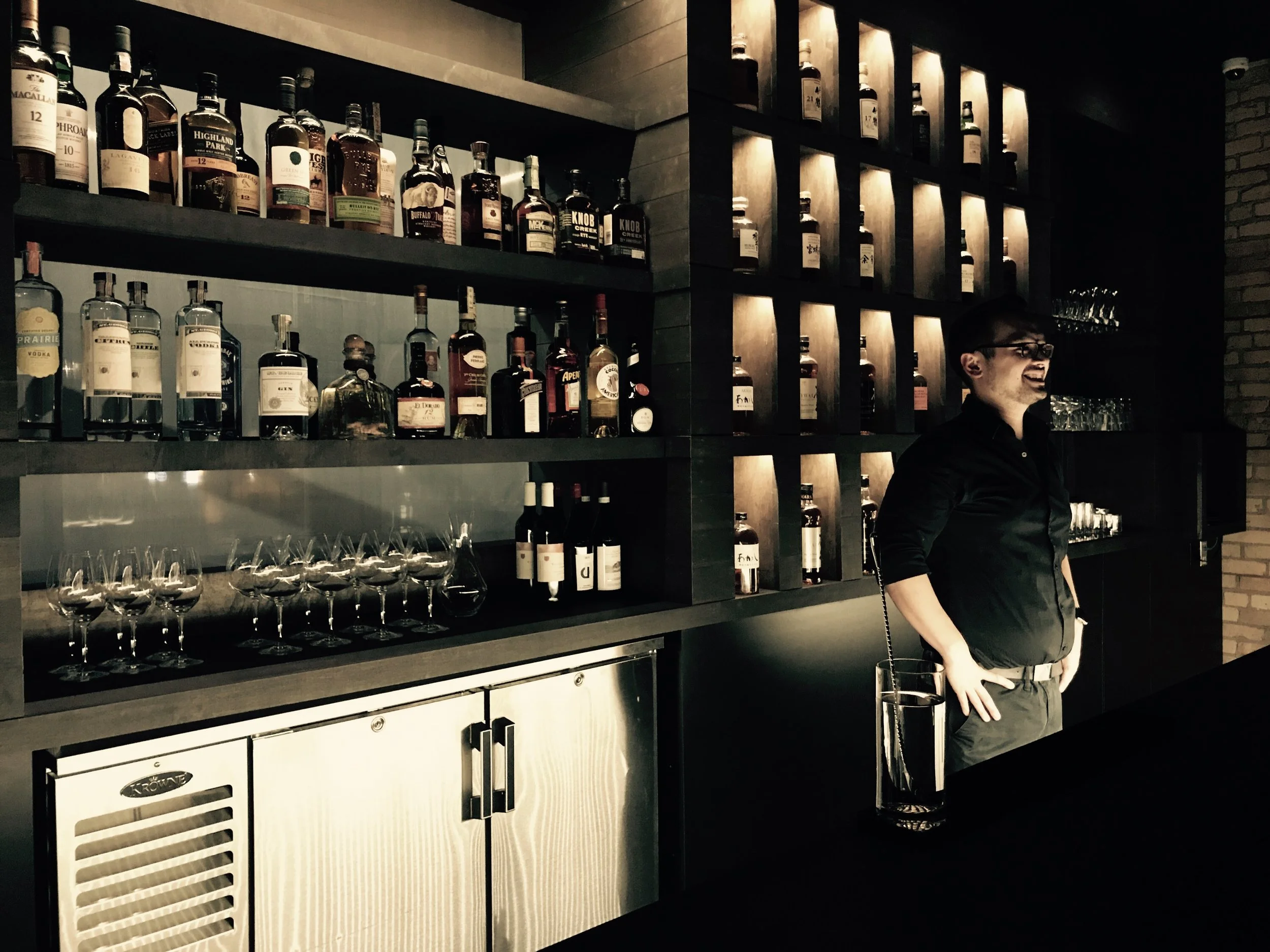 A bartender stands behind a bar with shelves of various liquor bottles, some illuminated. The bartender is smiling and wearing black. There are wine glasses on the counter, and the bar has a stainless steel refrigerator. The lighting is dim, creating
