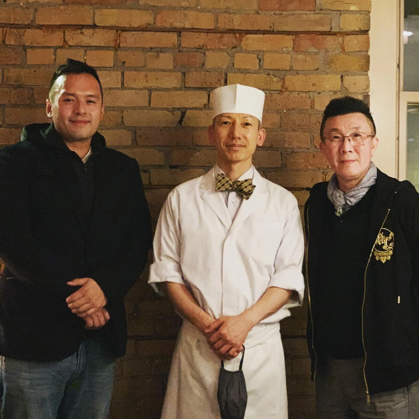 Three men standing in front of a brick wall, one dressed as a chef with a white hat and coat, the others in casual jackets.