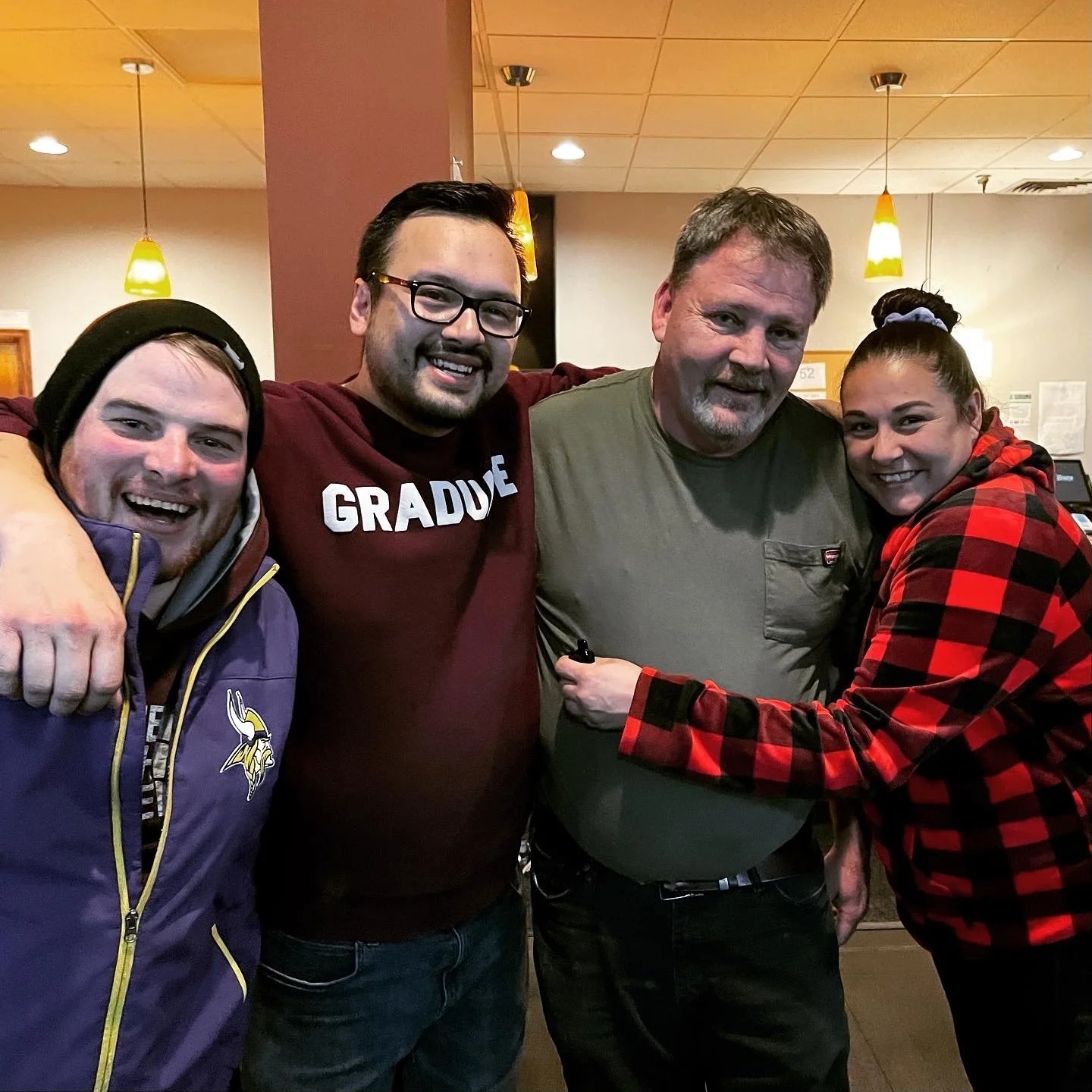 Group of four people smiling and hugging indoors, two men and one woman in a red and black checkered hoodie, one man in a green shirt, and another man in a purple Minnesota Vikings jacket.