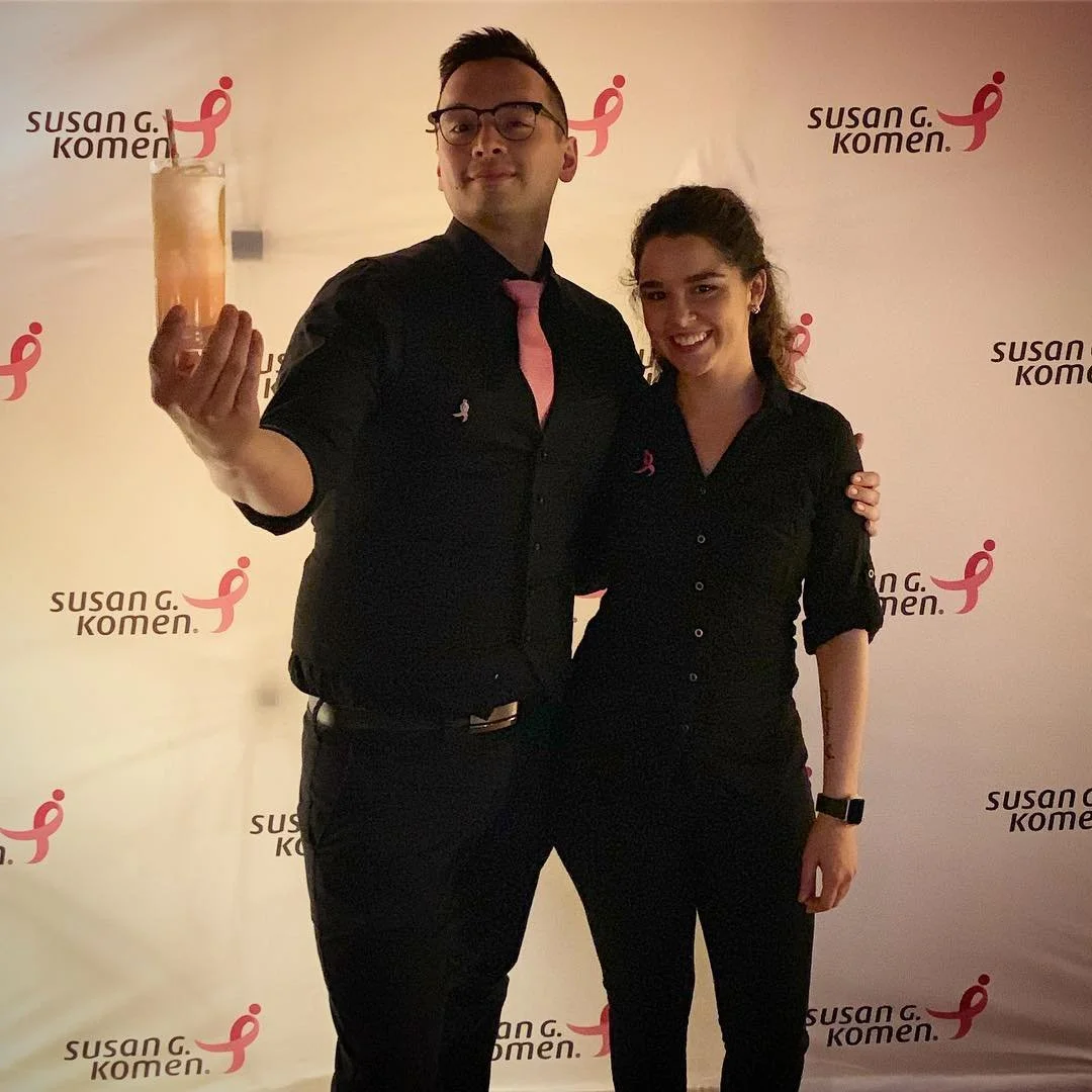 A man and woman stand together in front of a Susan G. Komen backdrop. The man holds a tall glass with a drink and a straw, and both are wearing black shirts with pink accents, smiling at the camera.