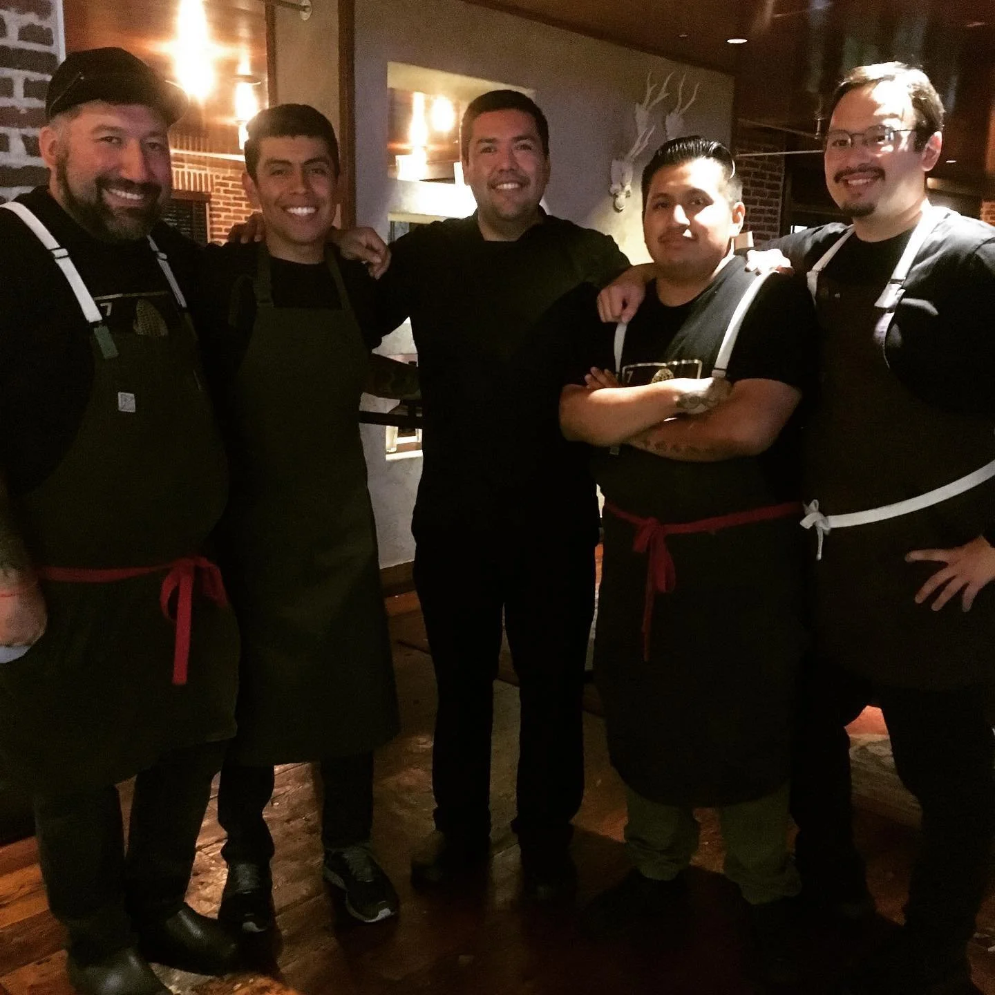 Group of five men in black shirts and aprons standing together in a restaurant, smiling for the camera.