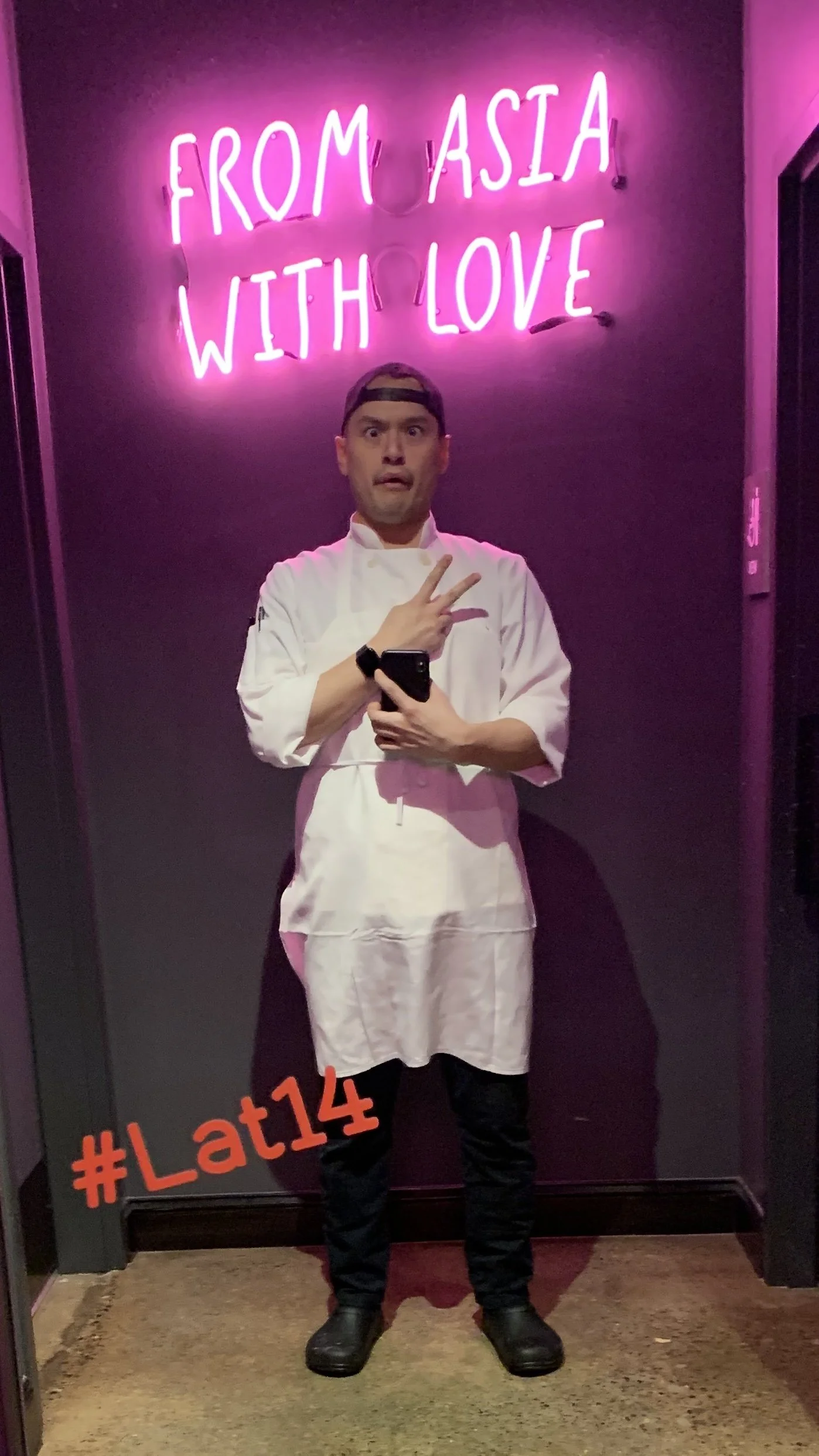 A man in a chef's coat taking a selfie in front of a neon pink sign that says 'From Asia with Love,' with a hashtag '#Lat14' in red at the bottom left.