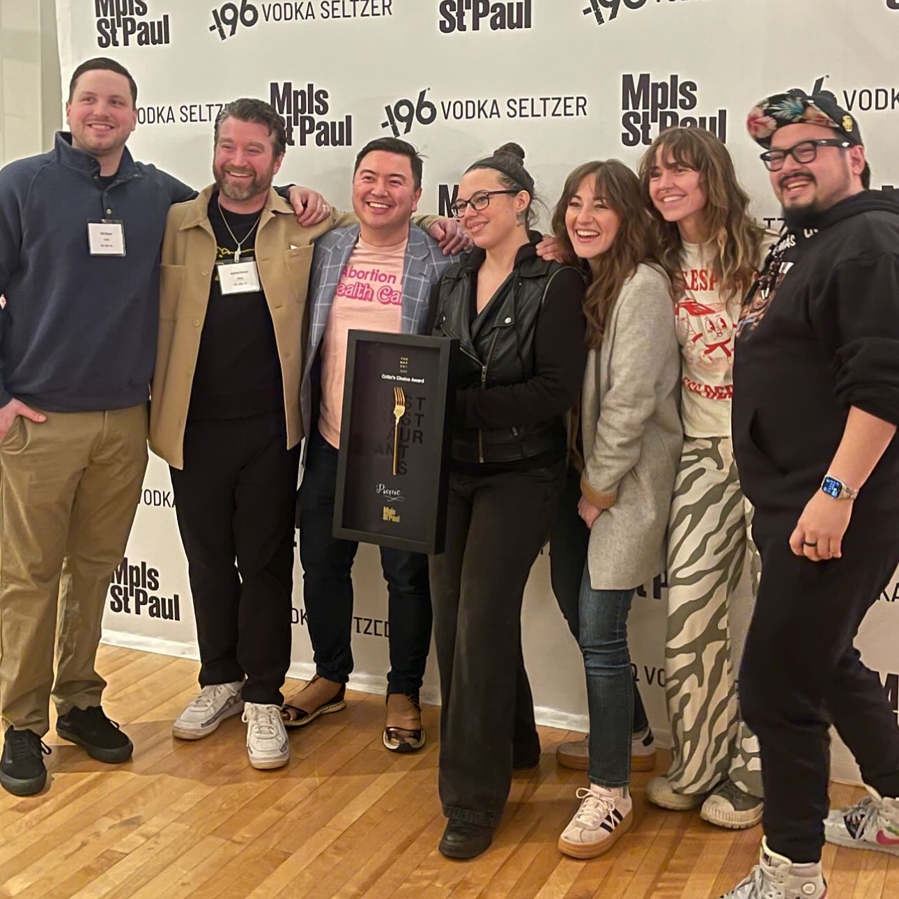 Group of seven people smiling and holding an award at an event, standing in front of a backdrop with text that reads 'Mpls St Paul' and 'Vodka Seltzer'.