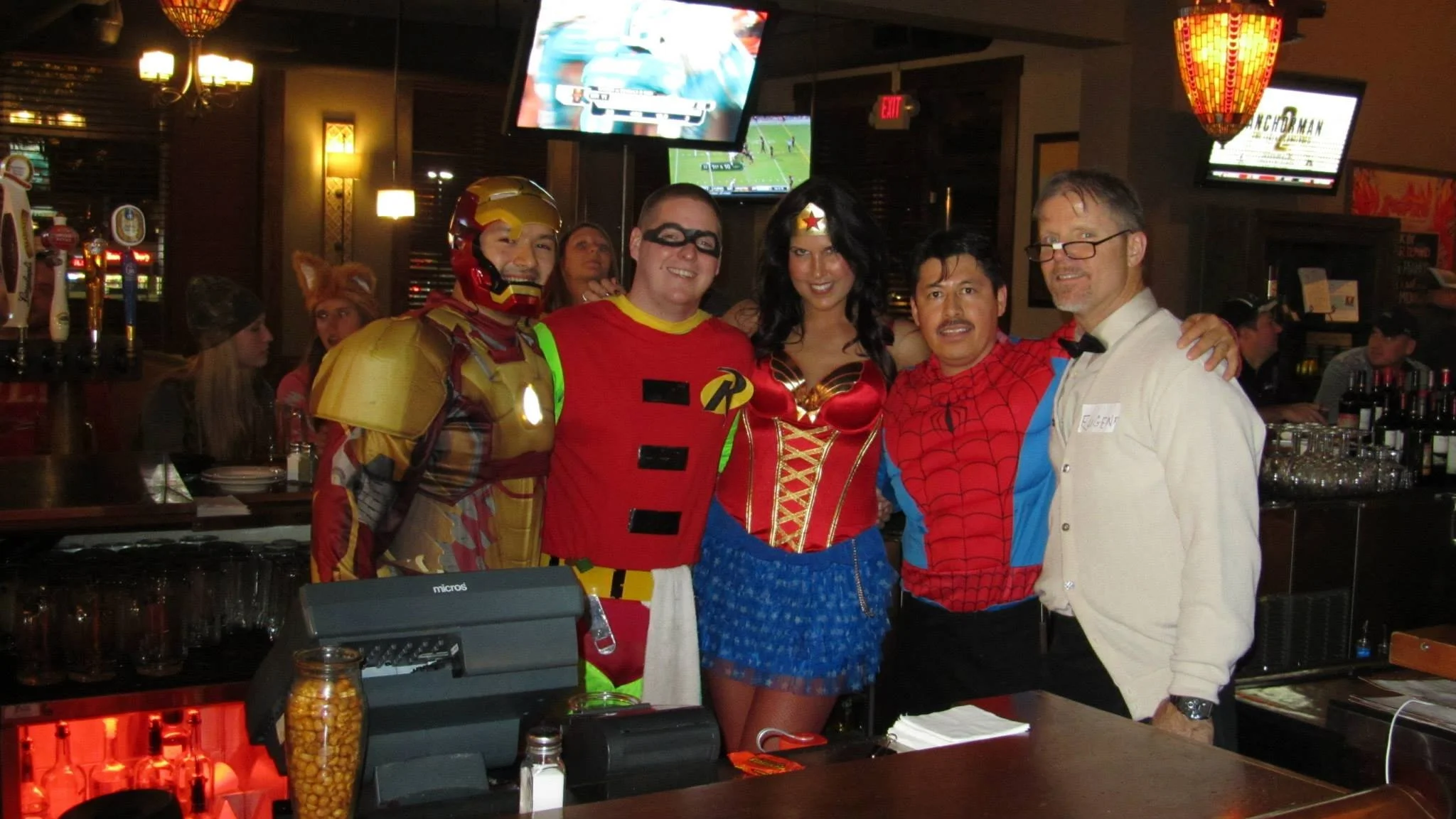 Group of six people in costumes at a bar, including one dressed as Iron Man, one as Robin, and one as Wonder Woman, with a man in a Spider-Man shirt, and a man in a white shirt and bowtie.
