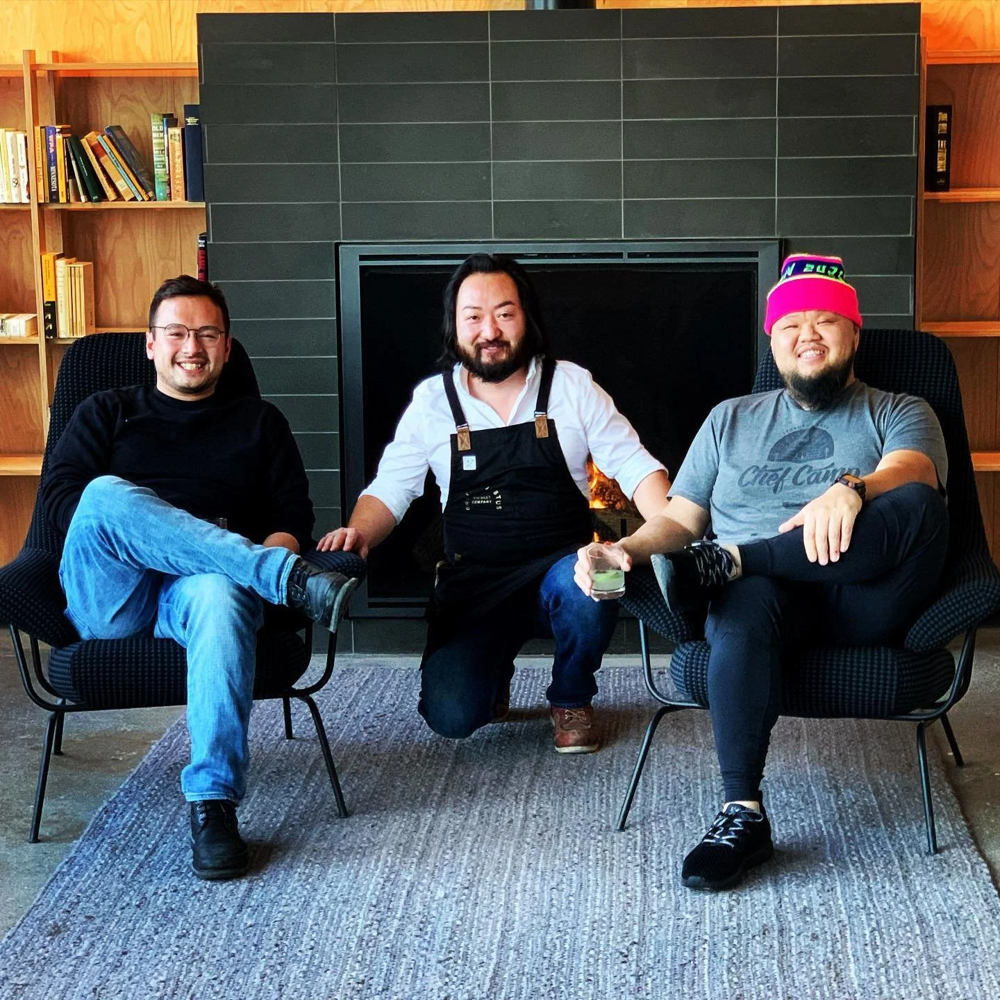Three men sitting in front of a fireplace, two on black chairs and one kneeling. They are smiling, holding drinks, and dressed casually with bookshelves in the background.