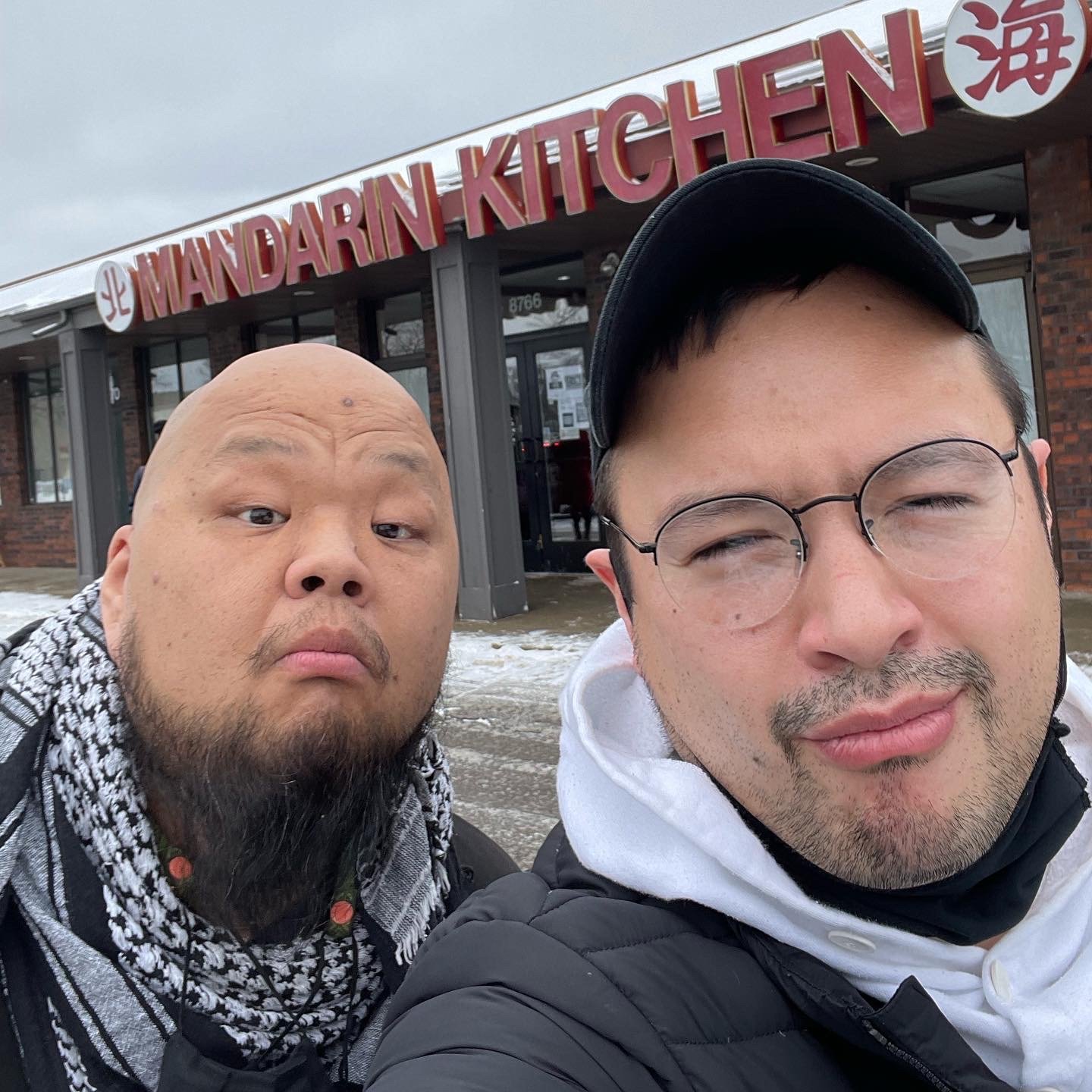 Two men taking a selfie outside a restaurant called Mandarin Kitchen. One man has a bald head and beard, the other has glasses, a cap, and facial hair. There is snow on the ground.