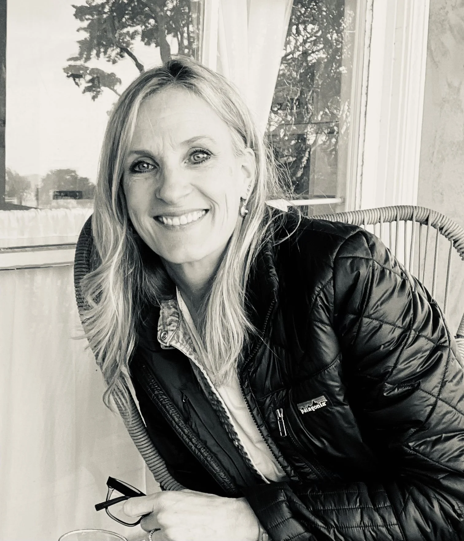 A woman with long blonde hair smiling, wearing a leather jacket and holding glasses, sitting in front of a window with curtains, in black and white.