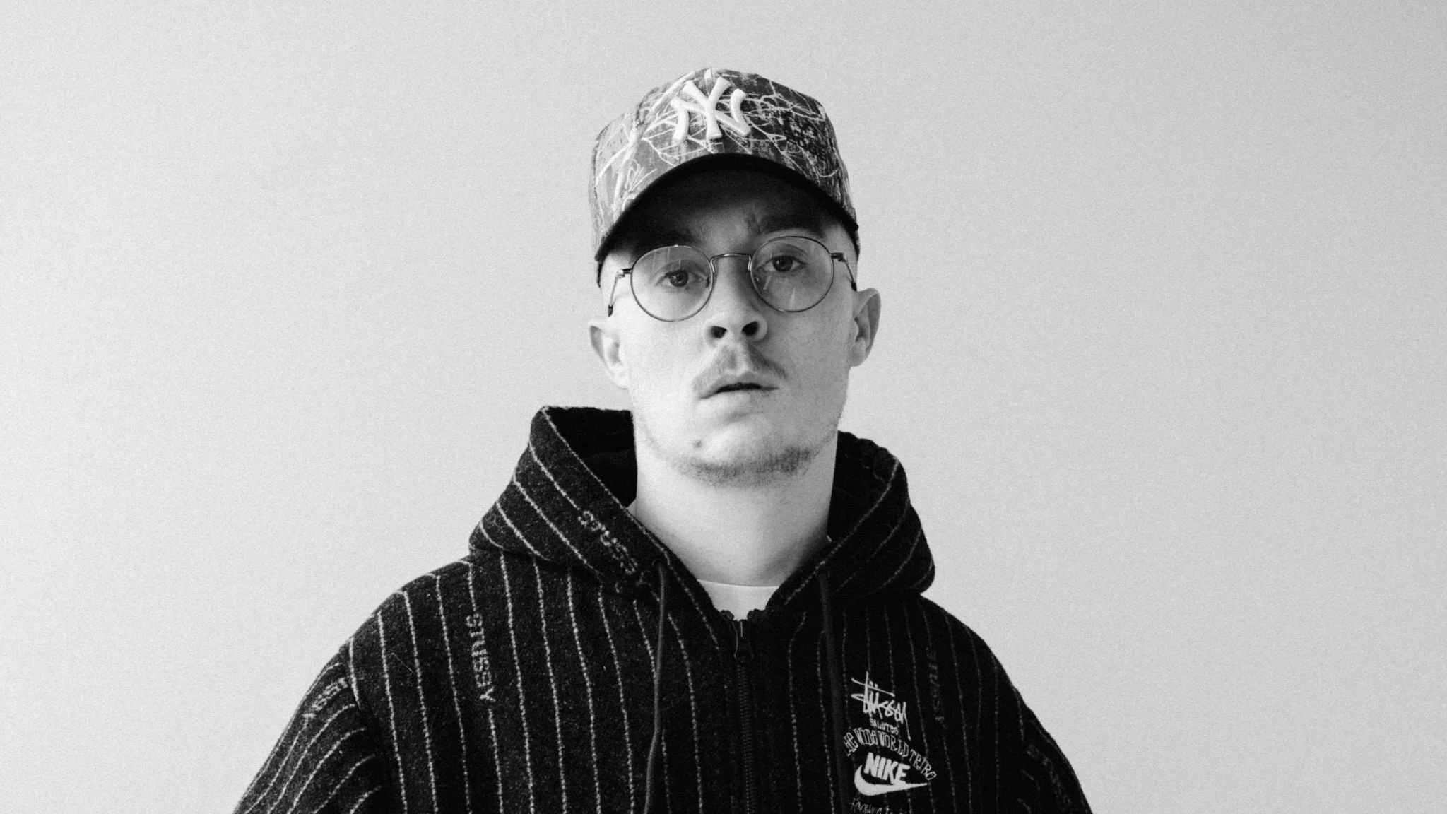 Black and white portrait of a young man wearing a baseball cap, glasses, and a Nike hoodie, against a plain background.
