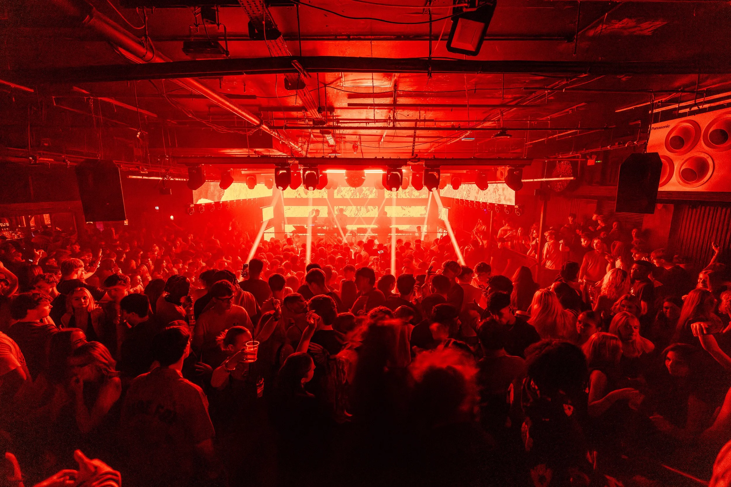 Crowded nightclub with people dancing and socializing under red lighting, with a DJ booth and stage in the background.
