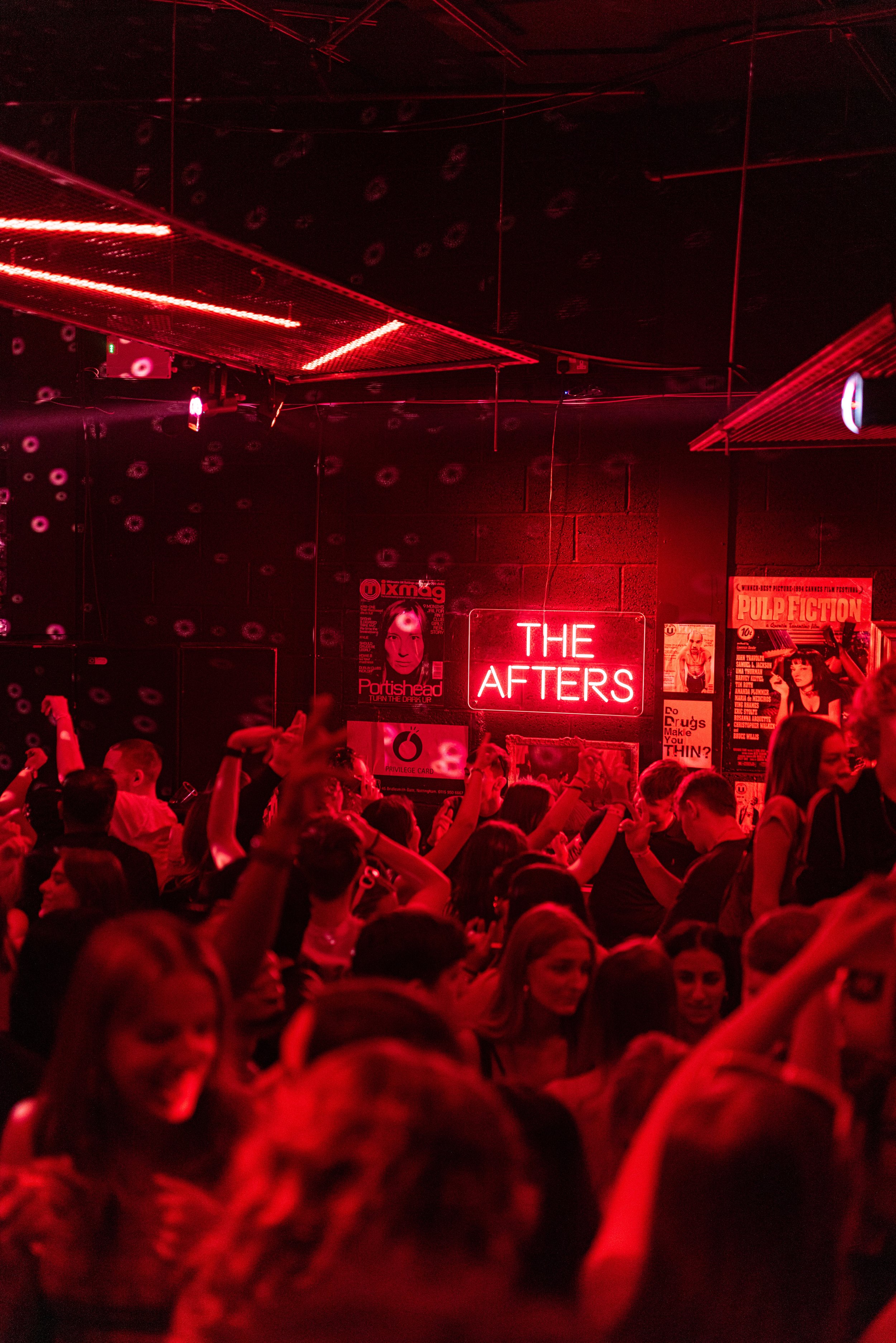 A crowded nightclub with red lighting, people dancing, and a neon sign that says 'THE AFTERS' on the wall.