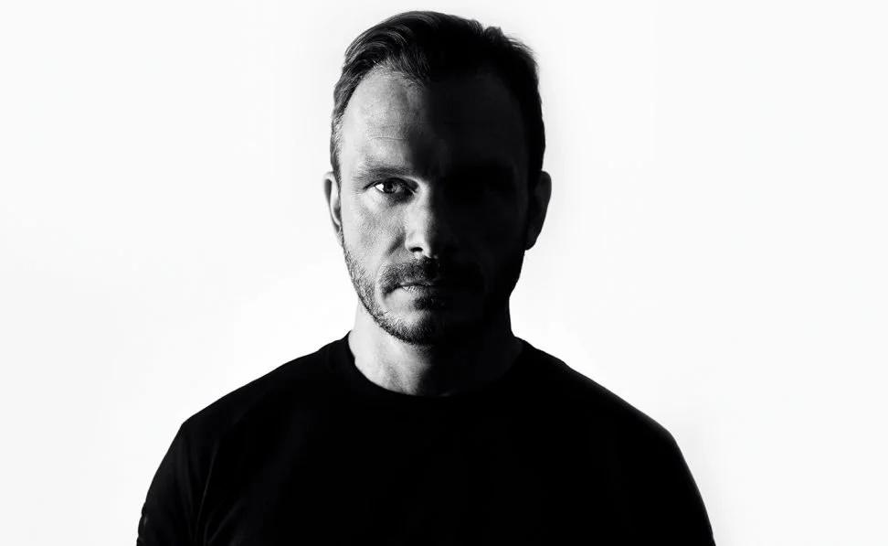 A black and white portrait of a man with short hair and a beard, wearing a dark shirt, against a plain white background.
