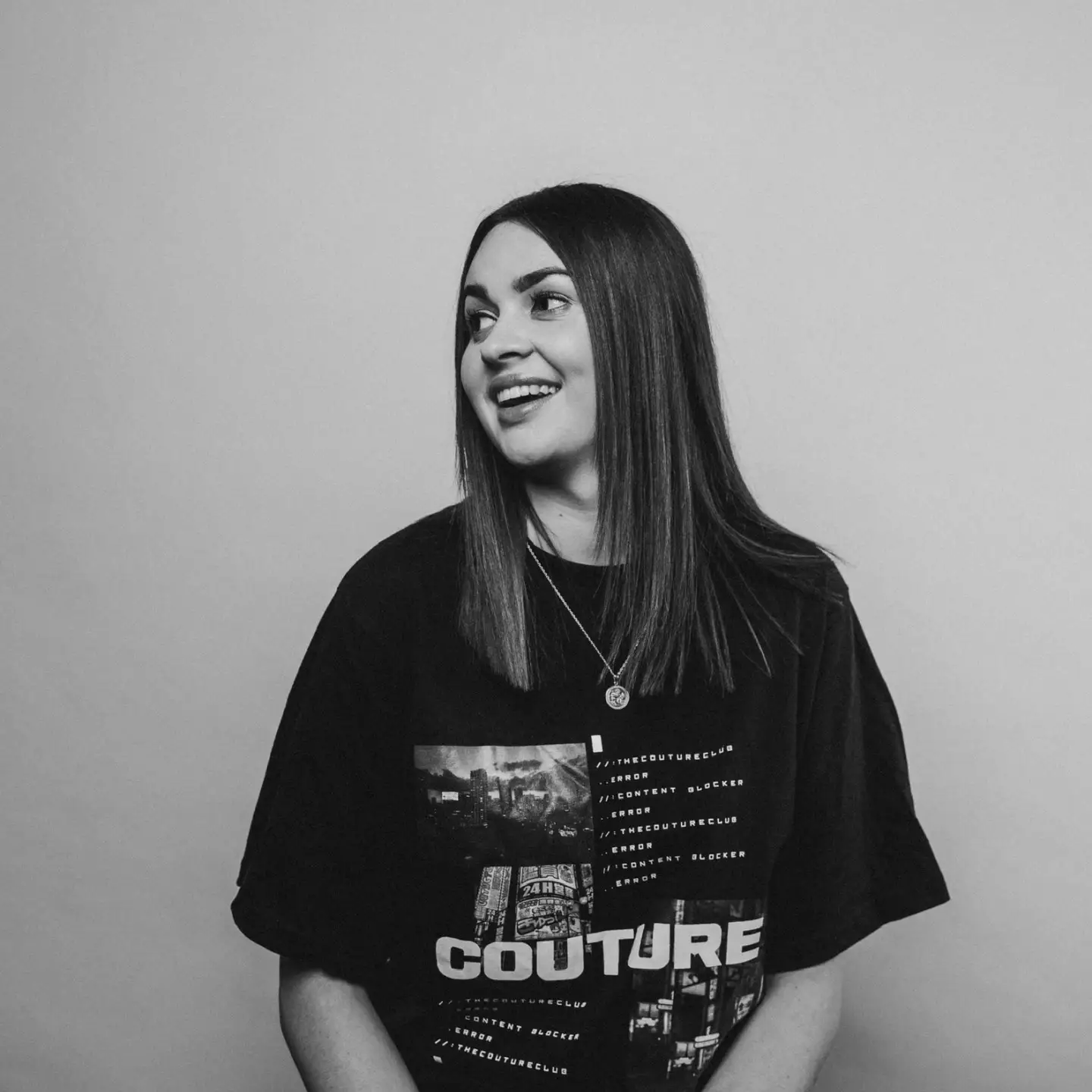 Black and white photo of a young woman with shoulder-length straight hair, wearing a graphic T-shirt and a necklace, smiling and looking to her left, standing against a plain wall.