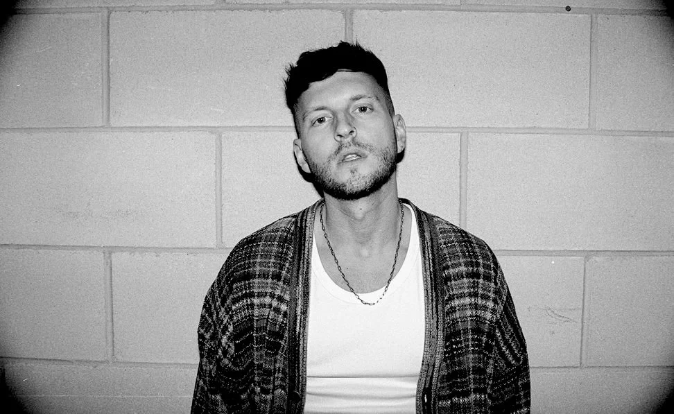 Black and white photo of a man with short hair and a beard standing against a brick wall, wearing a plaid cardigan over a white shirt and a chain necklace.