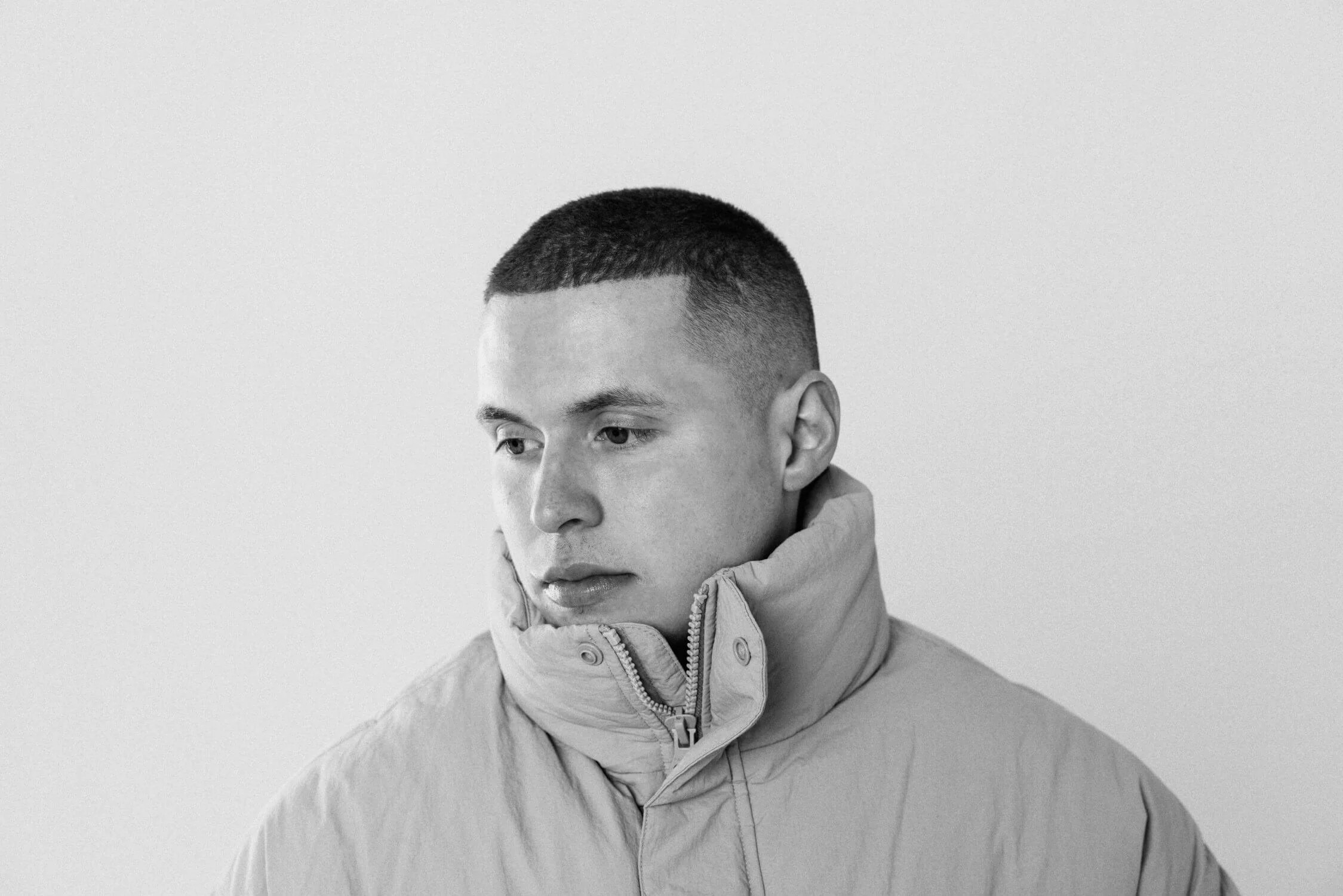 Black and white photo of a young man with short hair wearing a puffy jacket with the collar zipped up, looking to the side with a neutral expression.