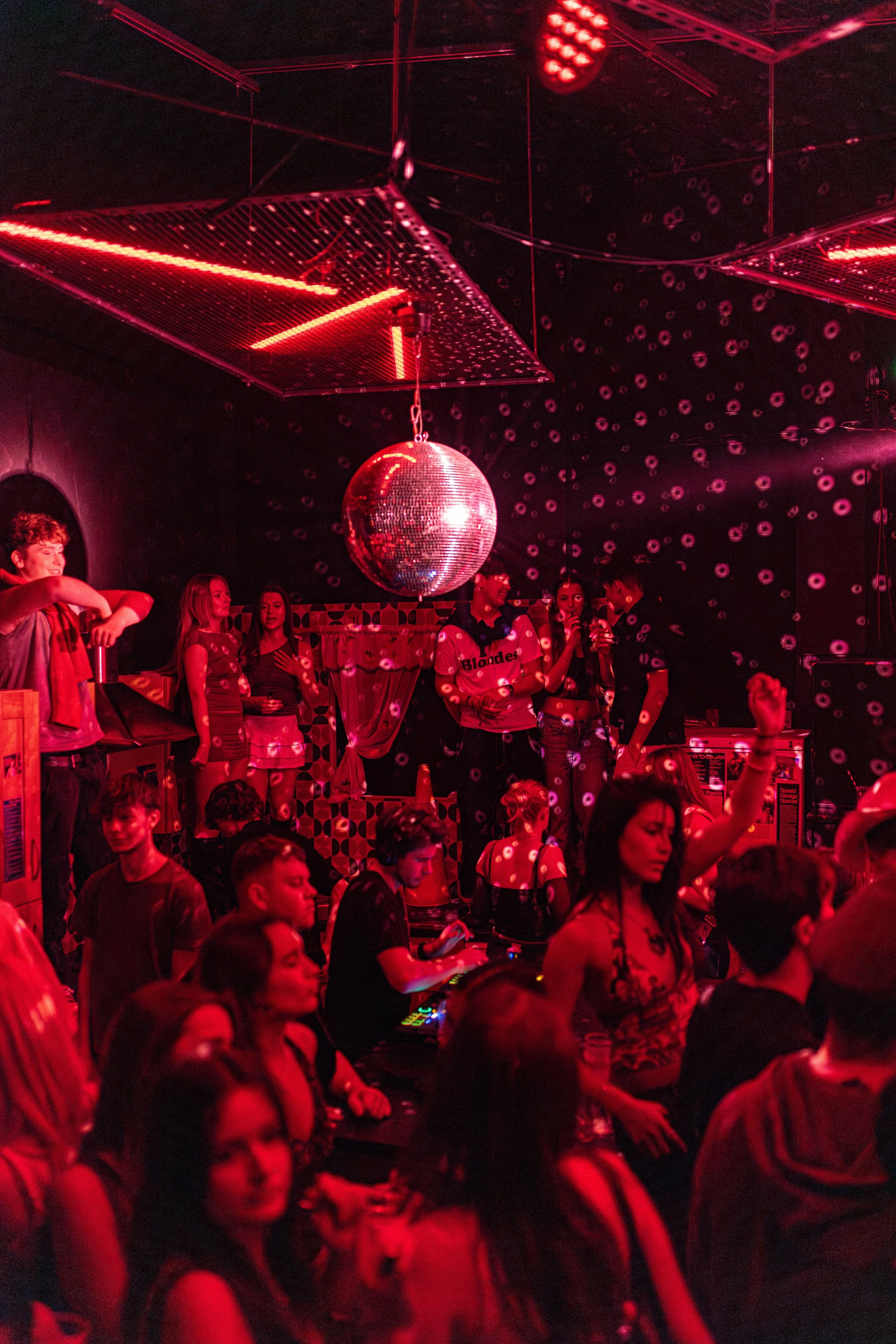 A crowded dance floor in a nightclub with red lighting, a disco ball hanging from the ceiling, and people dancing and socializing.