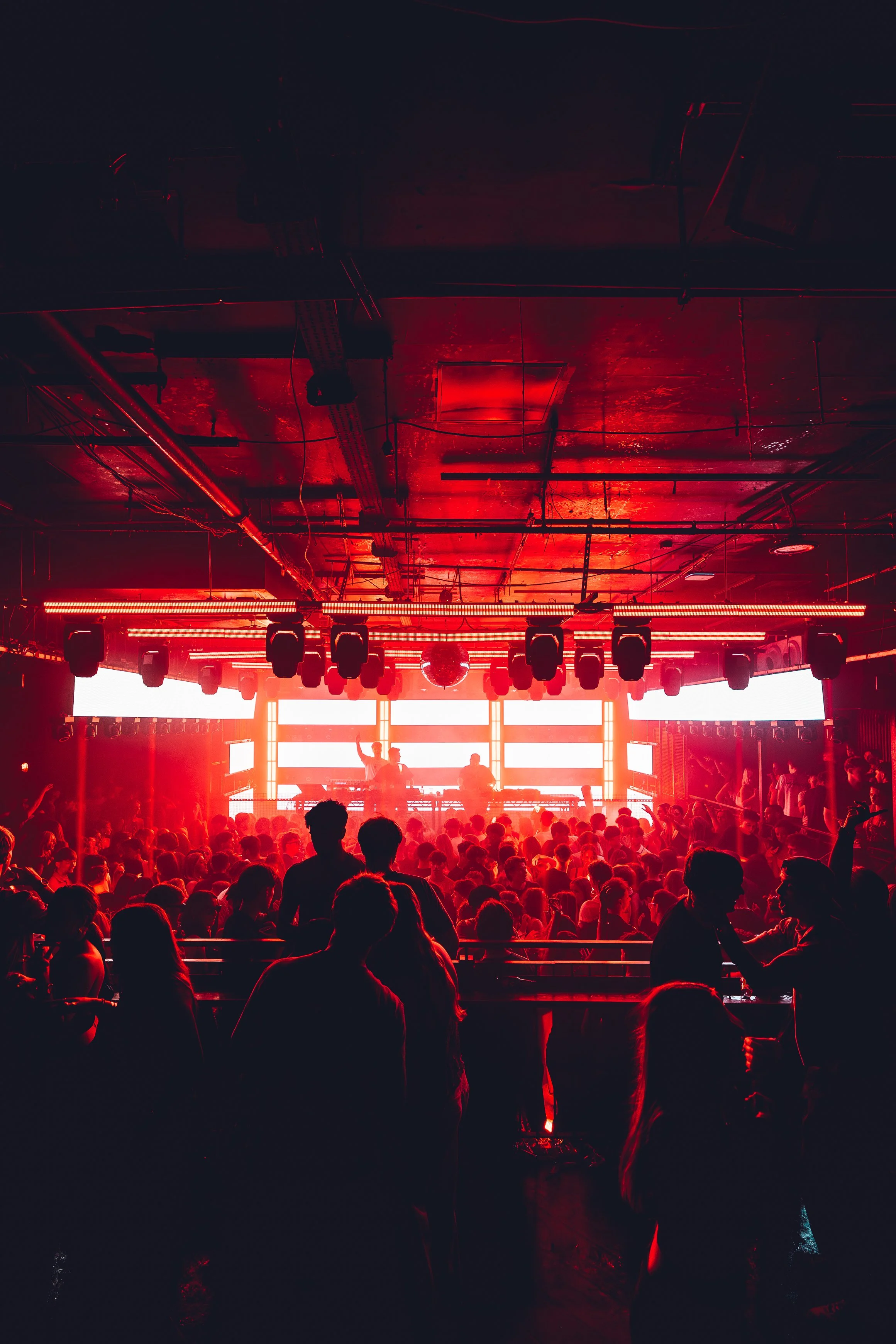 Crowd of people dancing and socializing at a concert or nightclub with red lighting and a DJ on stage.