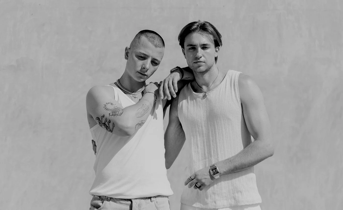 Two young men with lean builds standing close together, both wearing sleeveless tops, with one resting his arm on the other's shoulder, against a plain background in a black and white photo.