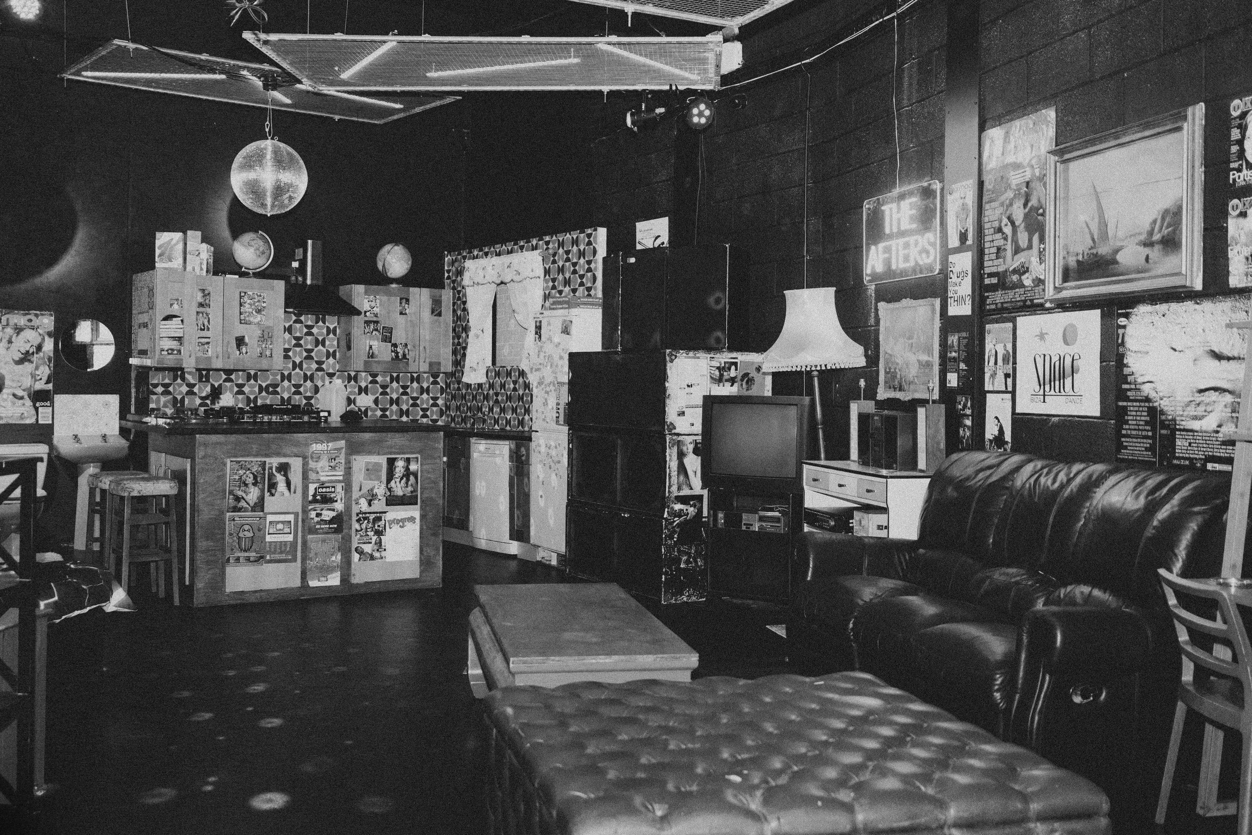 A vintage-style living room with a leather couch, an old TV, posters, a lamp, and a small bar area with stools, decorated with various light fixtures and artwork.