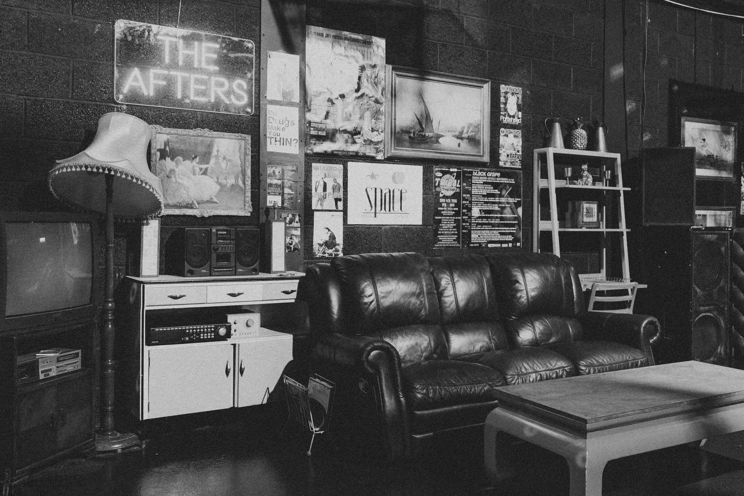A black and white photo of a cozy living room with a leather sofa, a vintage lamp, a TV, speakers, and a coffee table. The wall features various posters and artwork, including a sign that says "THE AFTERS."
