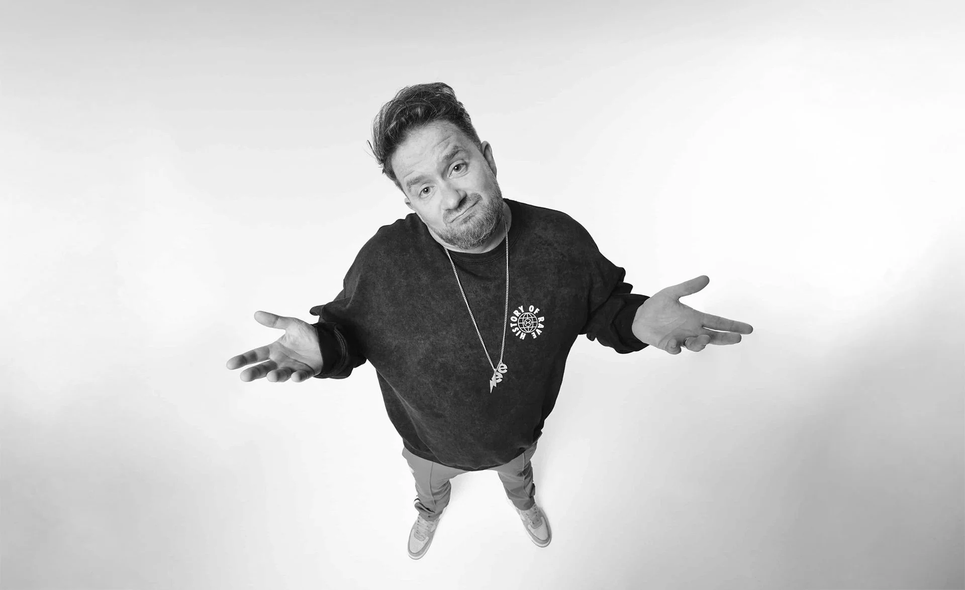 A man with a beard wearing a black sweatshirt, khaki pants, and sneakers, shrugging with palms facing up, against a plain light background.