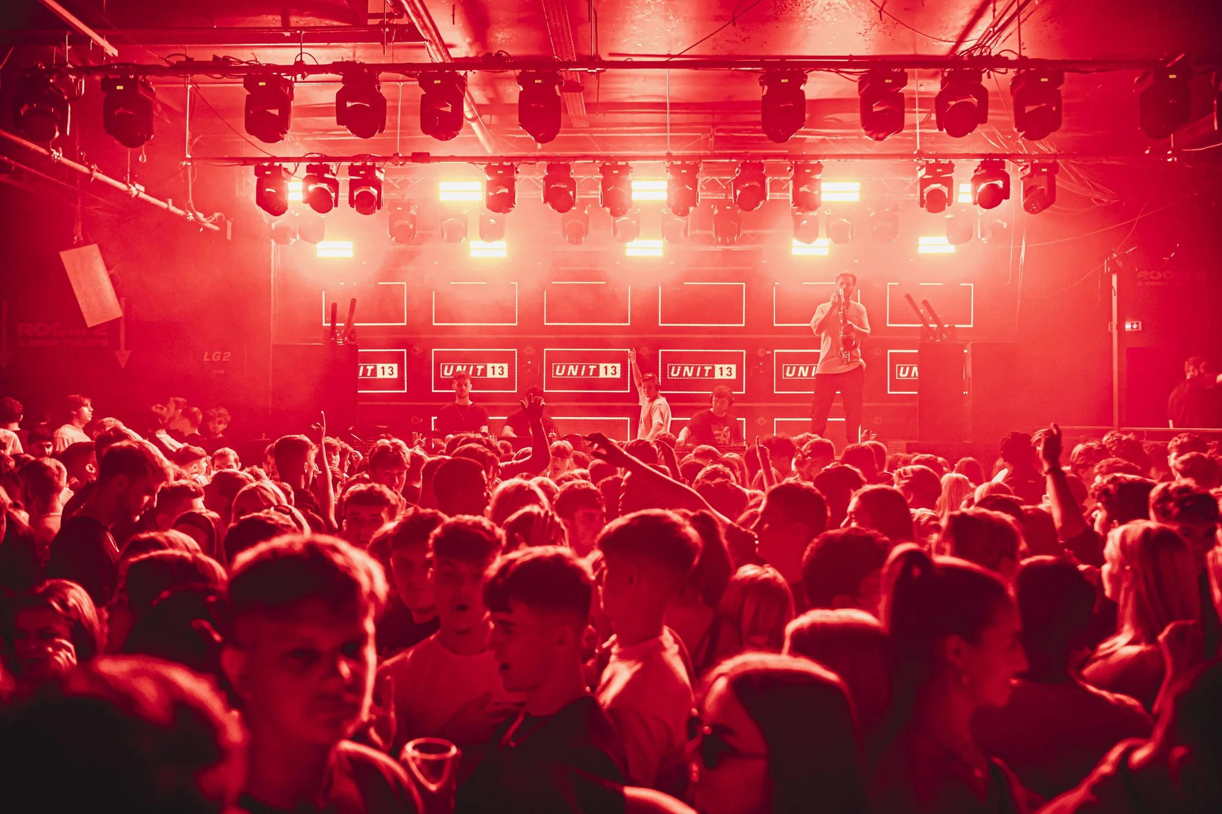 A crowded dance floor with people dancing in front of a stage lit with bright red lights and multiple screens displaying 'UNIT 13'.