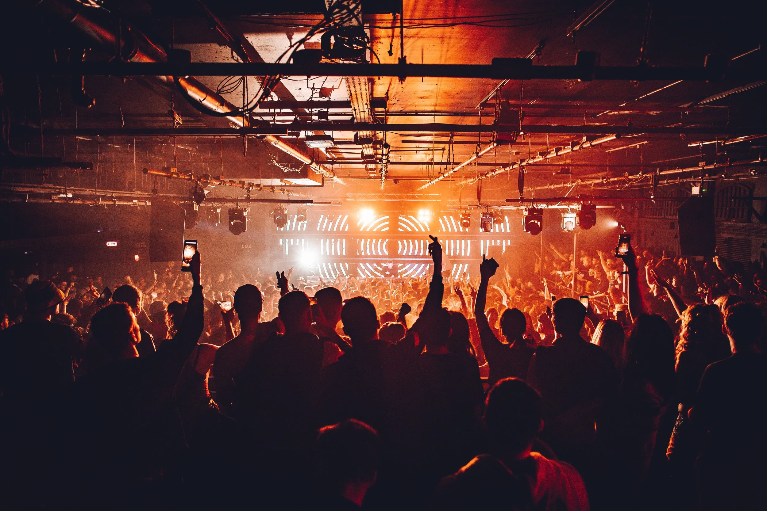 Crowd at an indoor concert or club with orange lighting and a digital neon display on stage.