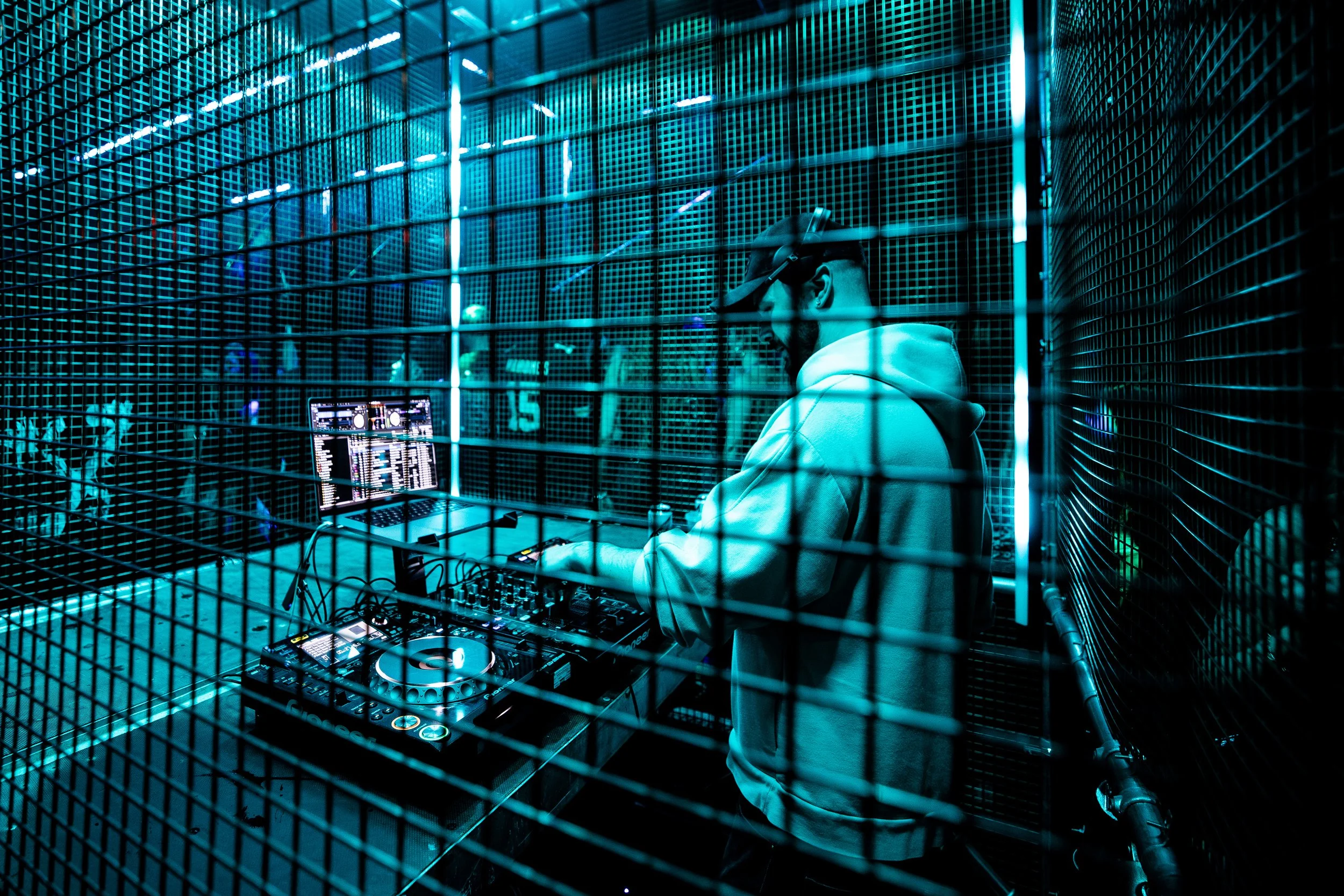 A DJ wearing a hoodie and virtual reality goggles operates a DJ controller behind metal fencing at a nightclub, with a laptop displaying music software nearby.