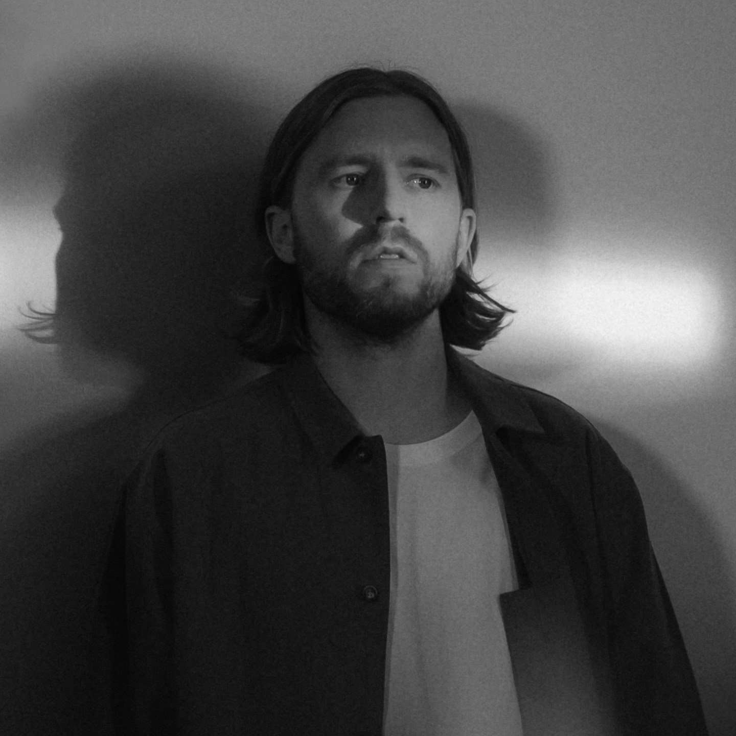 A black and white photo of a man with long hair and a beard, looking slightly upward with a serious expression, standing against a plain background.