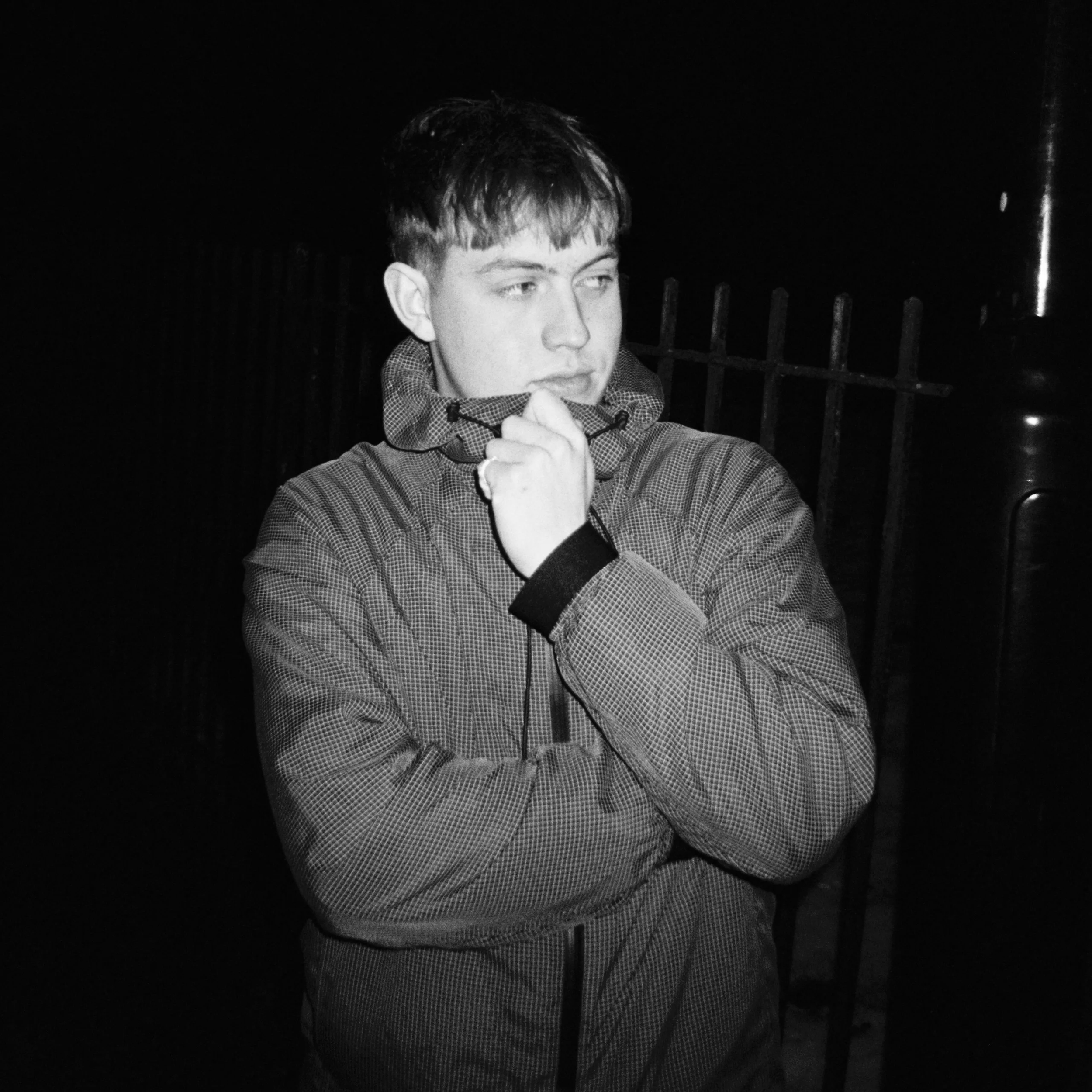 A young man in a checkered jacket standing outdoors at night, looking to the side with his hand near his face.