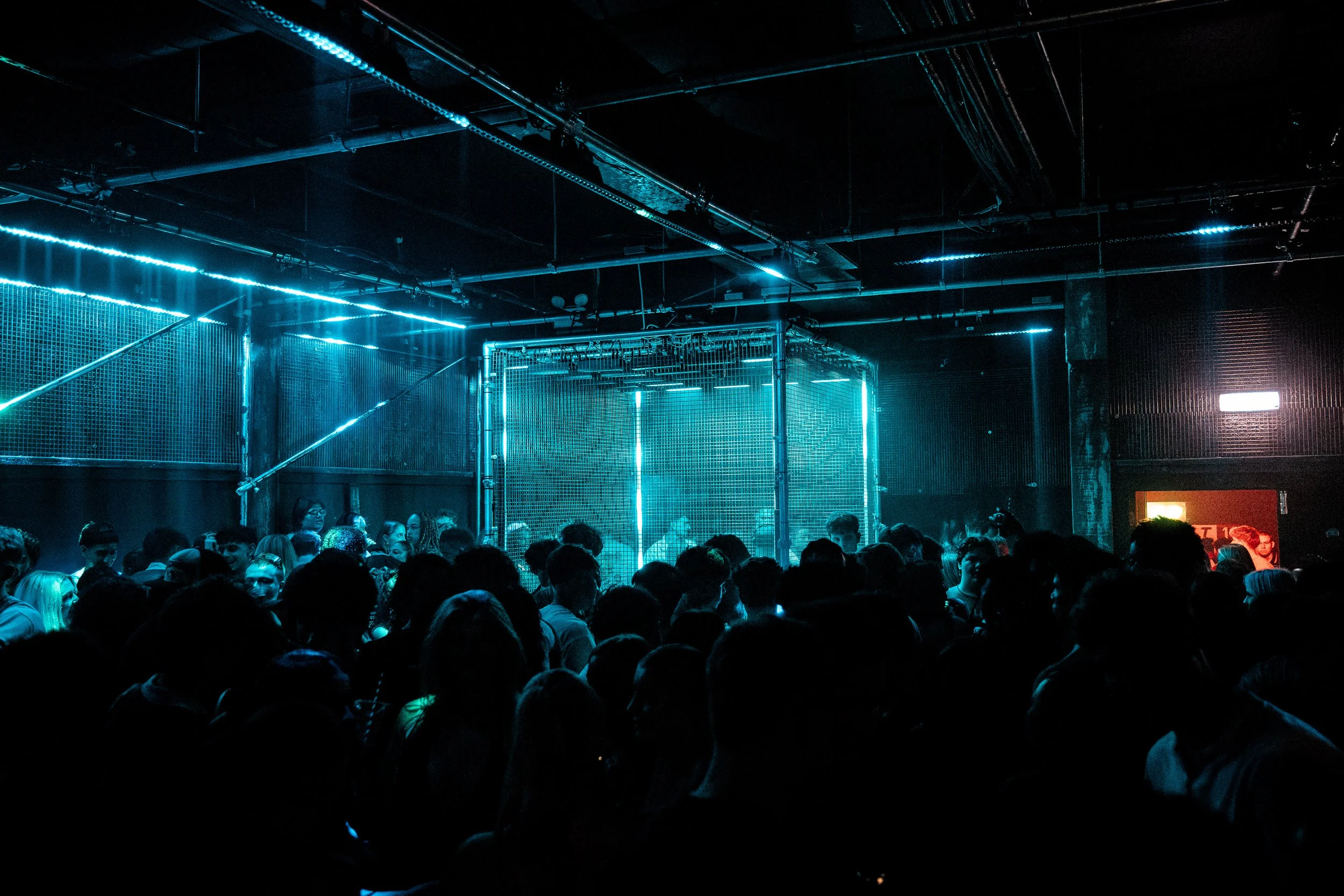Dark indoor concert venue with neon blue lighting, a crowd of people, and a metal cage stage in the middle.