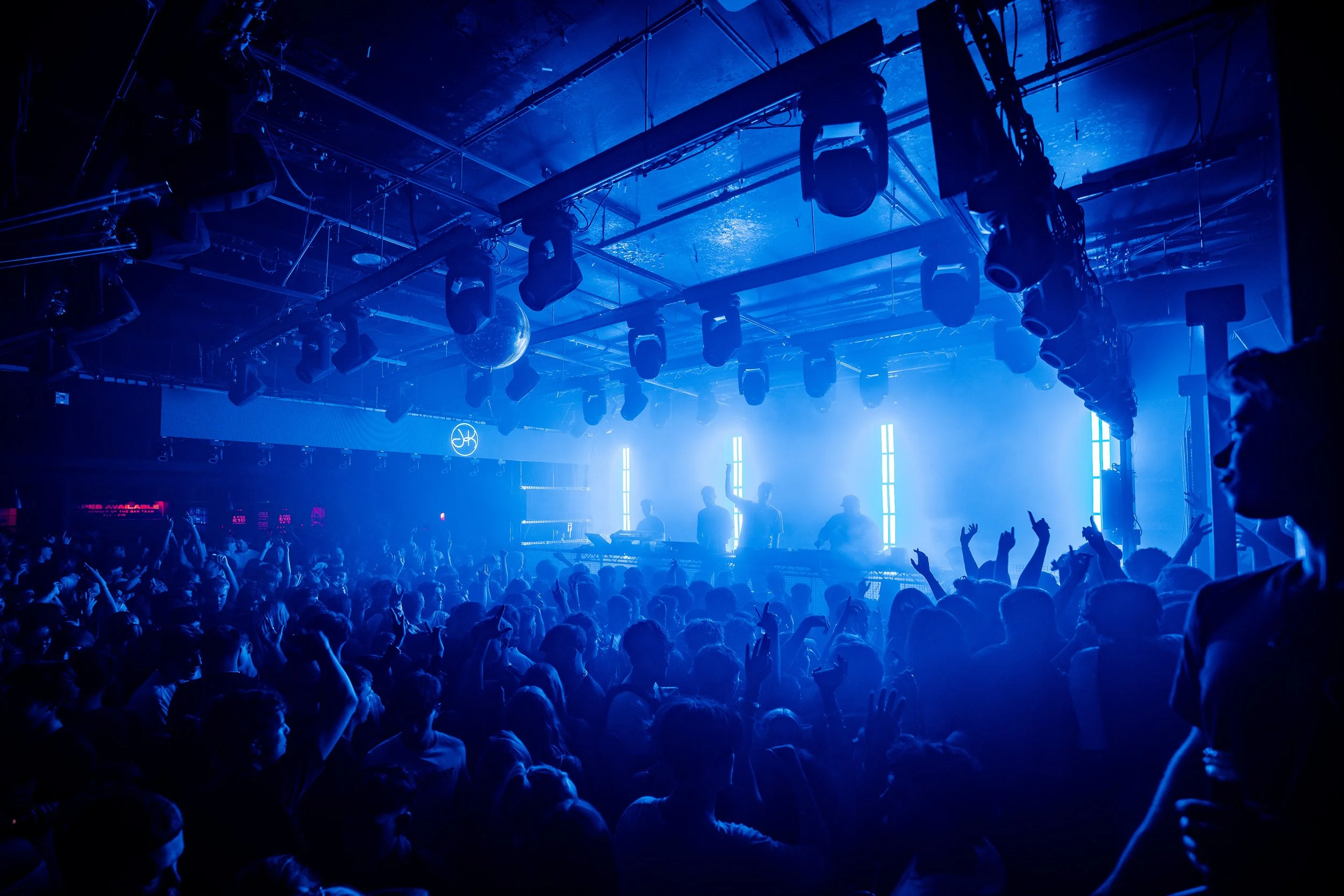 A crowded concert or nightclub scene with blue lighting, silhouettes of people dancing and raising their hands, a DJ or performers on stage, and various lighting equipment hanging from the ceiling.