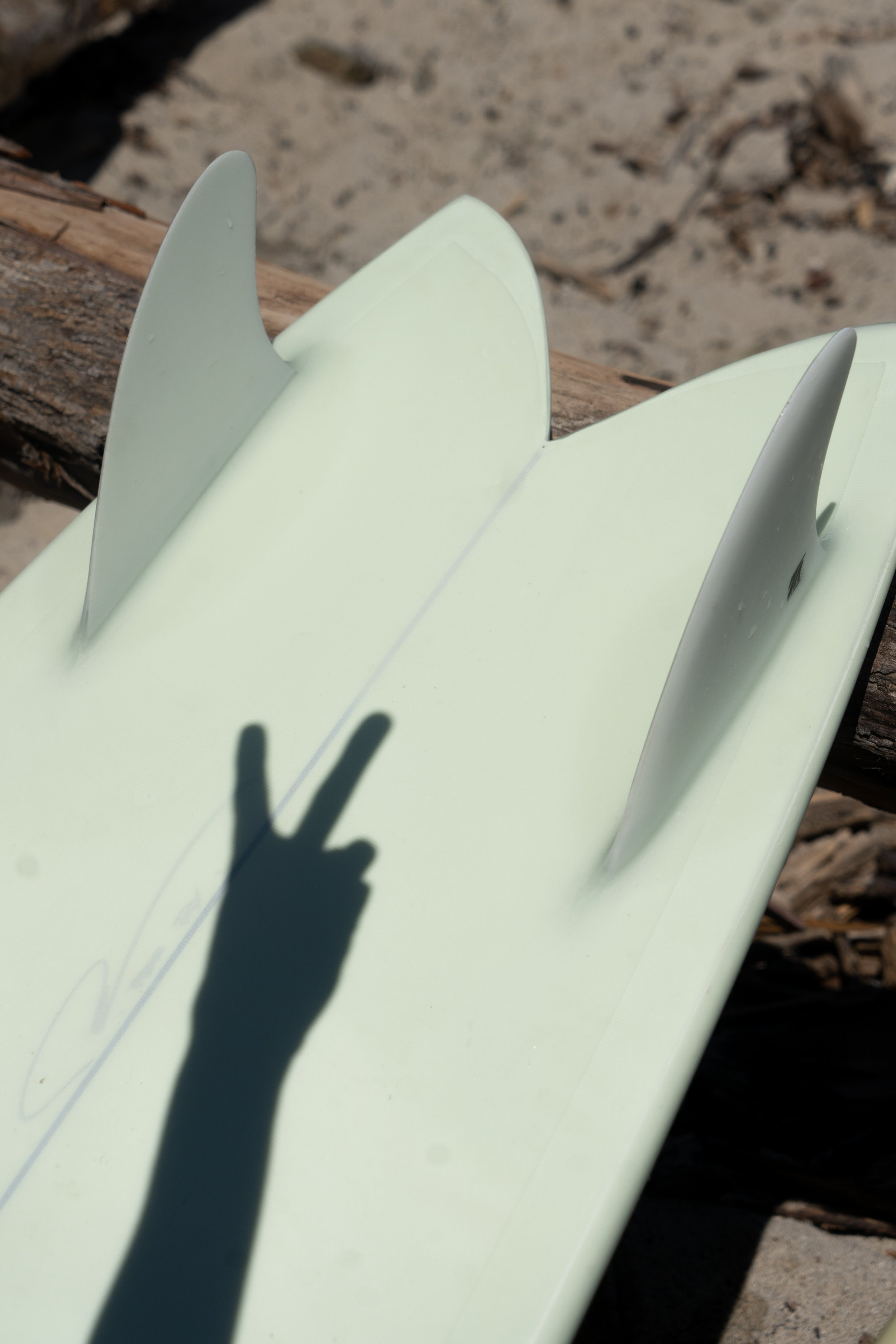 A surfboard with white fins resting on a piece of driftwood on the beach, with the shadow of a person making a peace sign on the surfboard's surface.