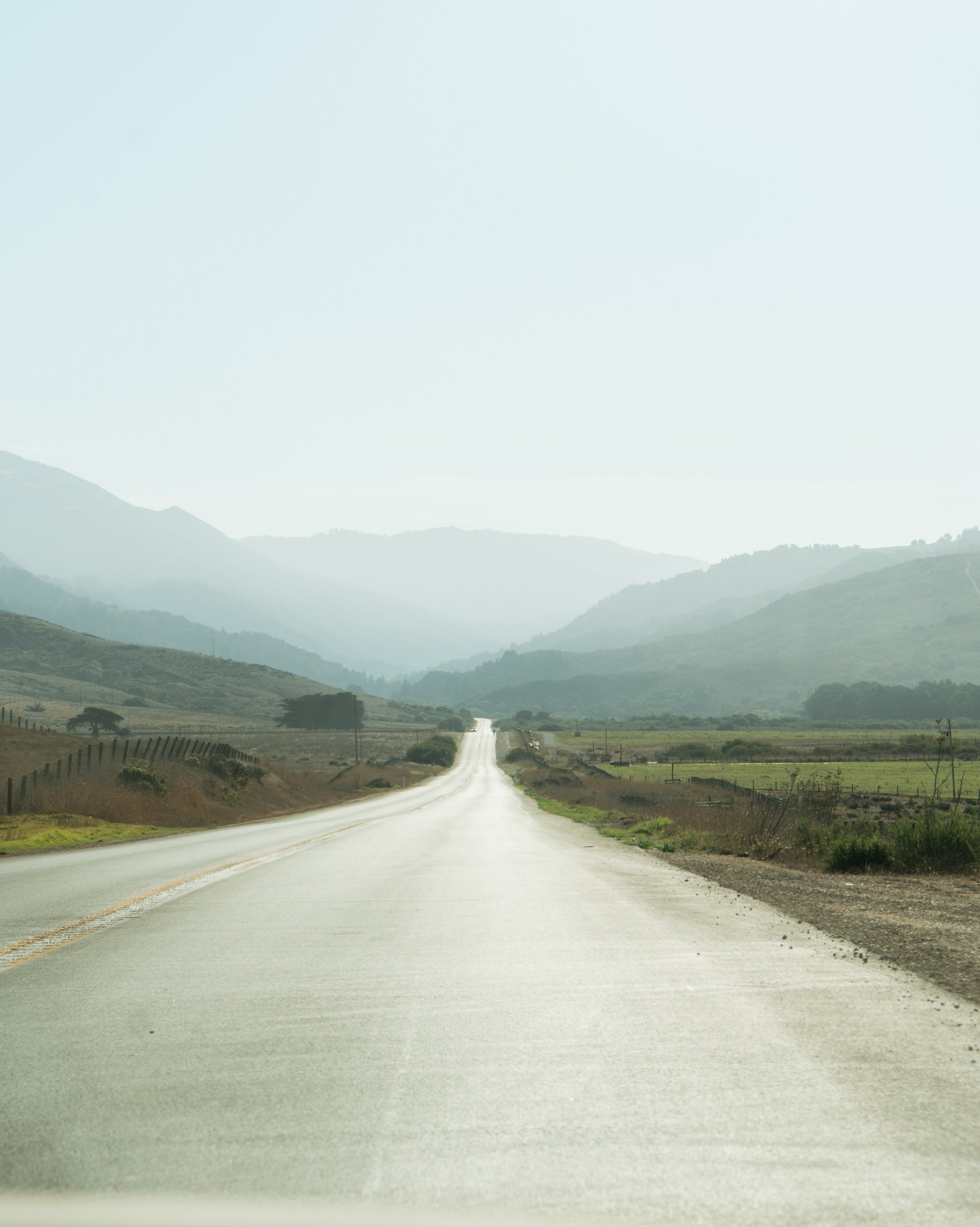 Empty rural road stretching into the distance with hills and mountains in the background, under a hazy sky.