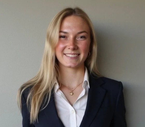 Young woman with long blonde hair, wearing a white shirt and blazer, smiling at the camera against a plain wall.